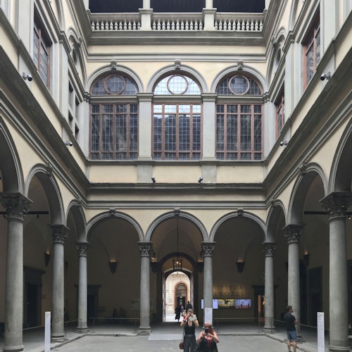 Two women stand in a museum, looking at a religious painting featuring the Virgin Mary with child and saints, framed by a gold triptych.