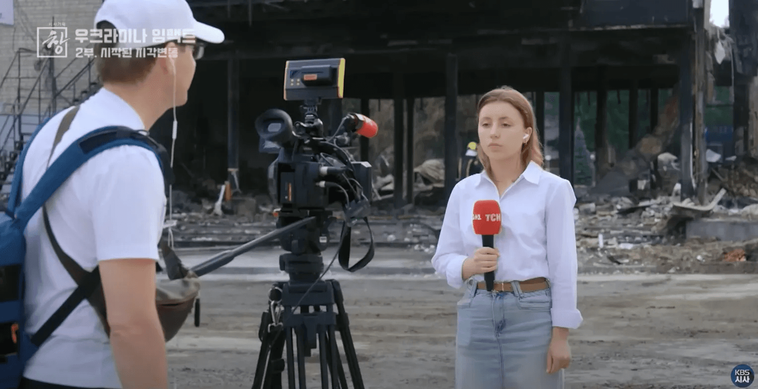 우크라이나 공습 피해 현장에서 리포트하는 우크라이나 방송 기자. 배경에 파괴된 건물 잔해가 보이며, 취재진이 촬영 중인 모습.