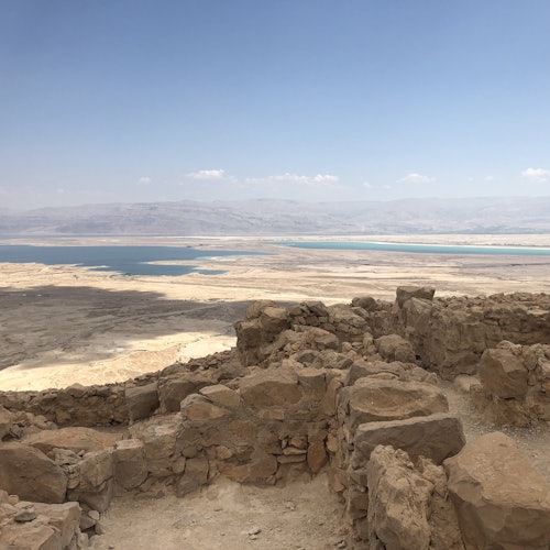 View of ancient stone ruins with a vast desert landscape and a large body of water in the background under a clear blue sky.