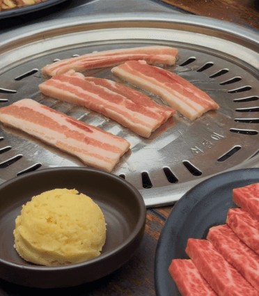 Two people serving food from wooden platters at an outdoor dining setup. A table is set with a white tablecloth.