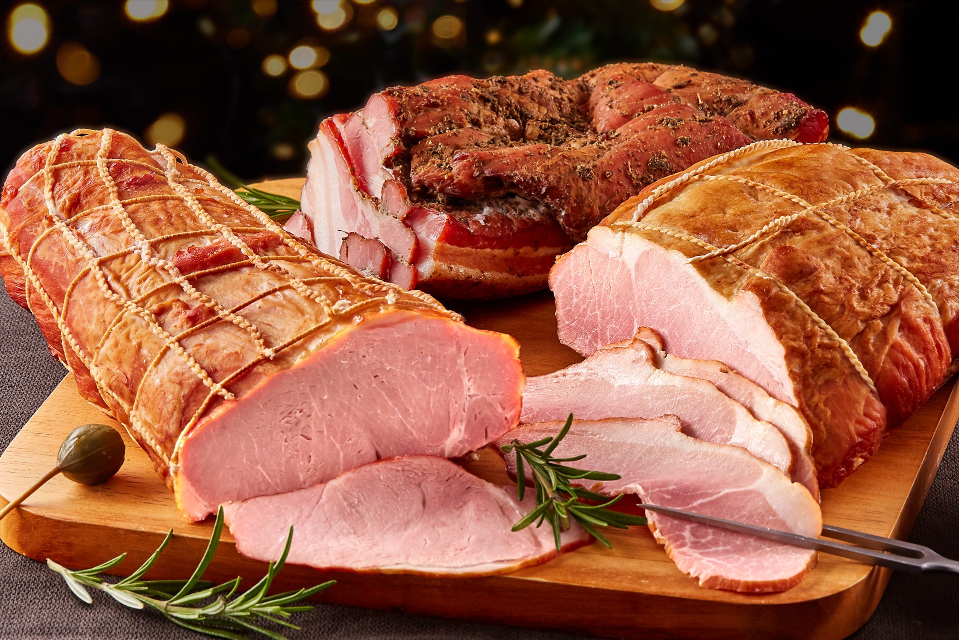 A wooden cutting board displaying three types of sliced, cured meats garnished with fresh rosemary and a caper, set against a background of warm, festive lights.