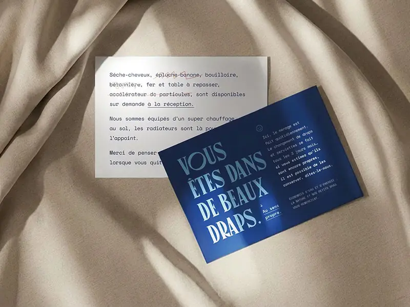 Deux cartes de communication créées pour l’Hôtel Outa : une carte blanche avec une liste de services barrés de façon humoristique et une carte bleue avec le message typographique “Vous êtes dans de beaux draps”, posées sur un drap beige.