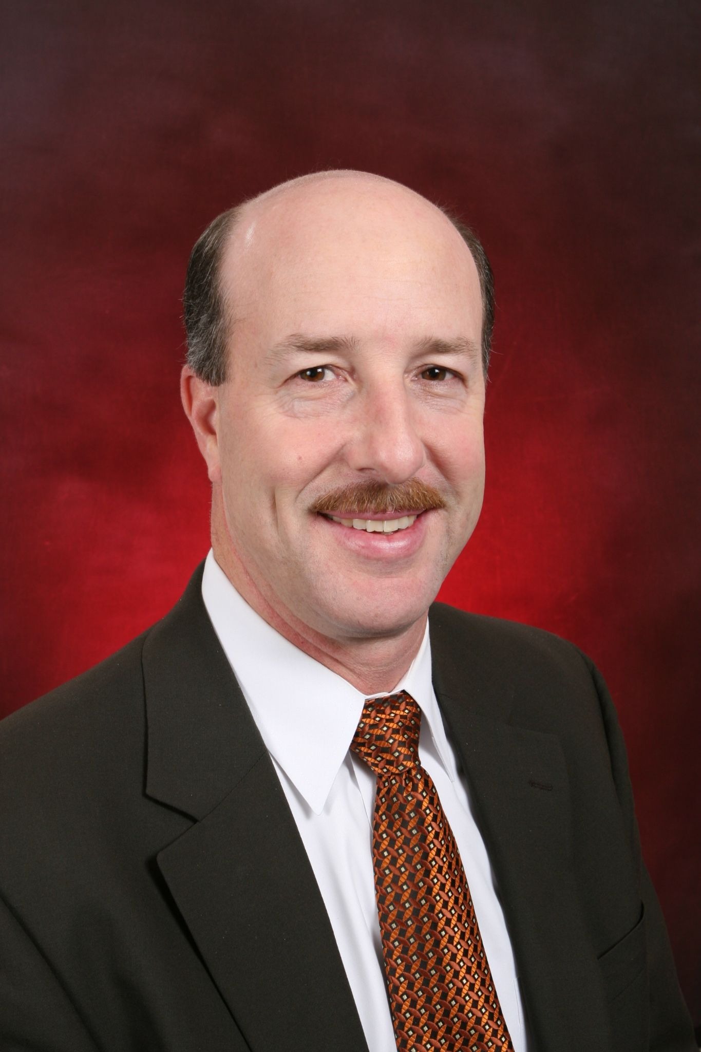 Professional headshot of David Bissonette in suit and patterned tie against red studio background.
