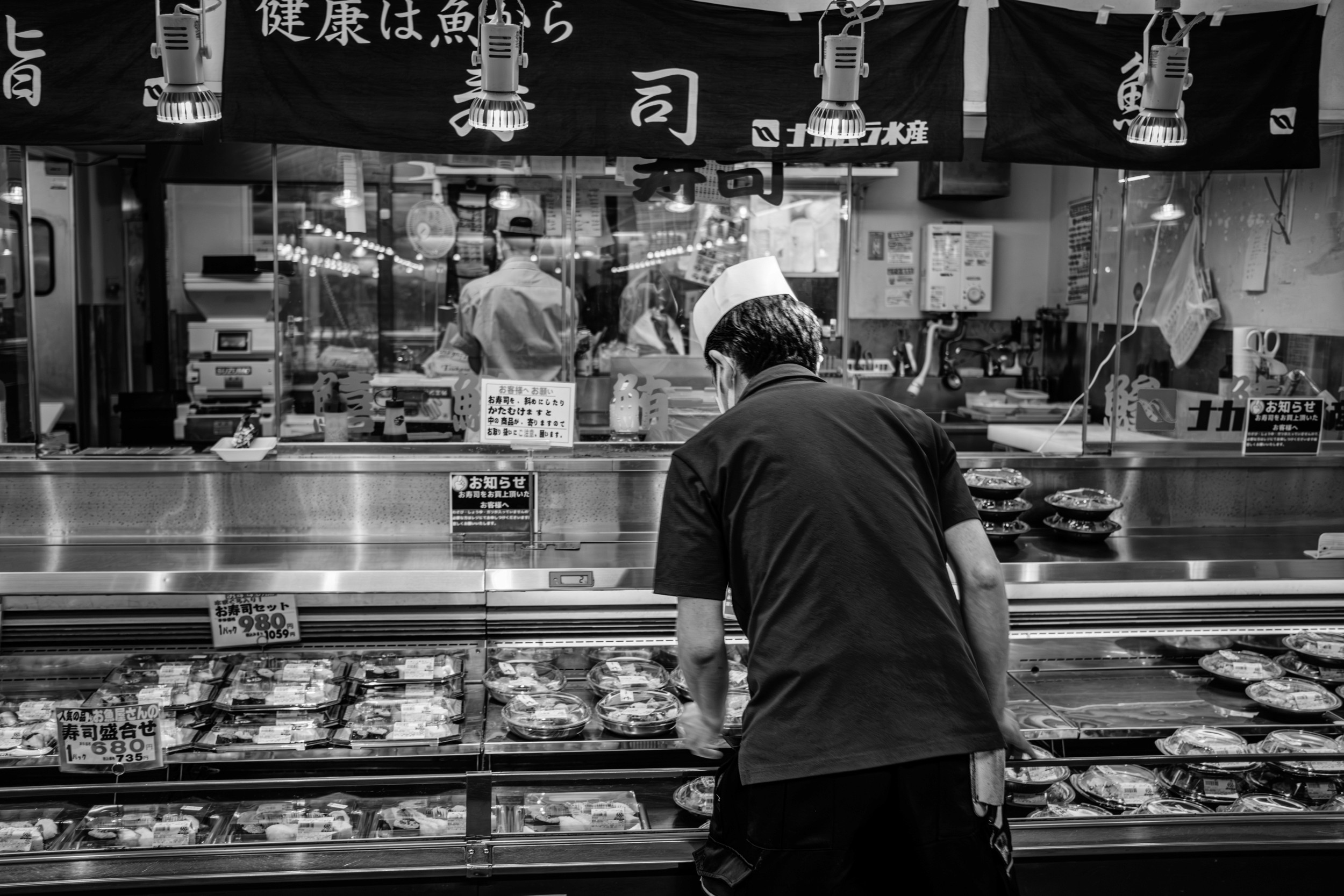 Black and white photograph of a sushi worker leaning over a refrigerated display arranging packaged sushi trays.