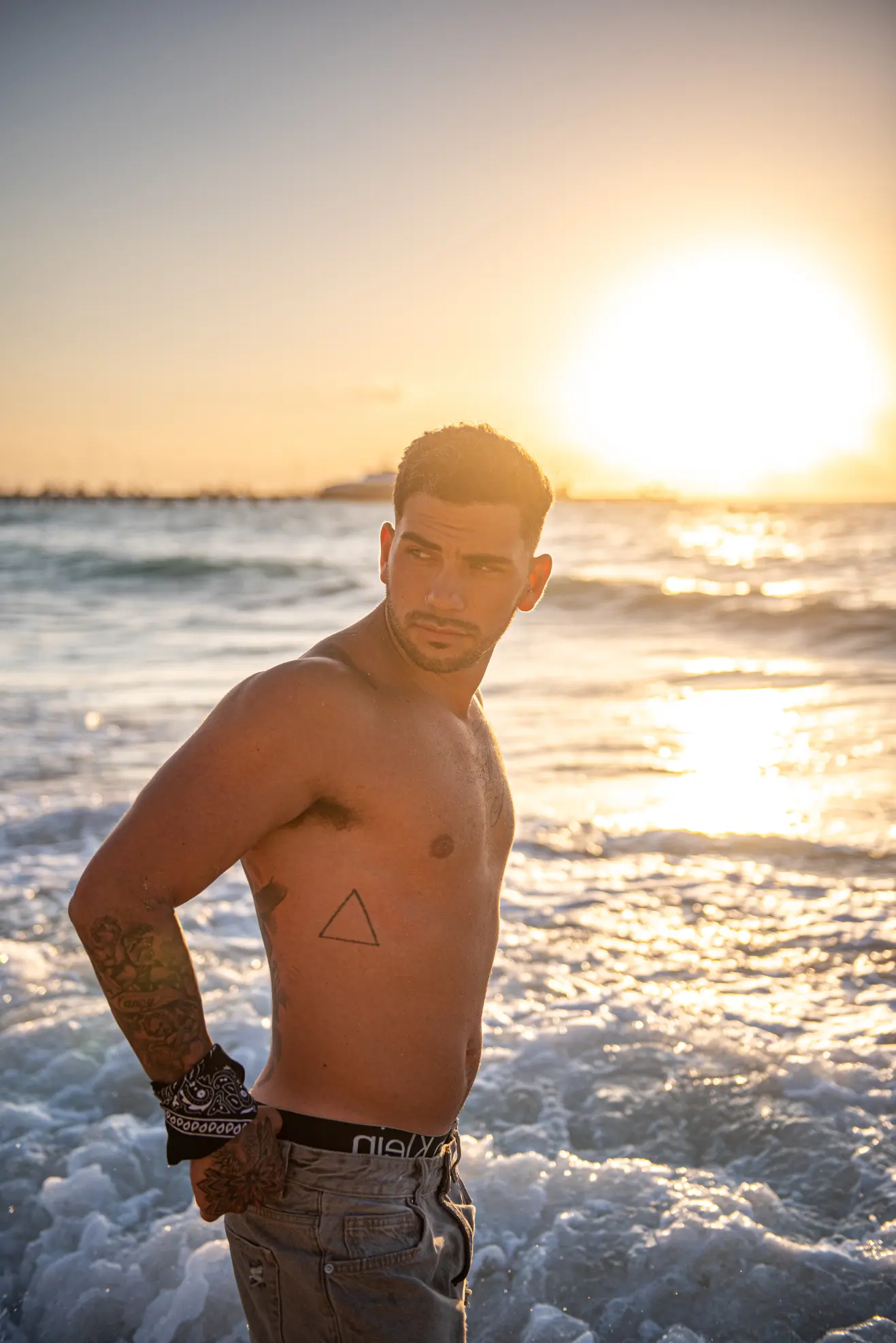 Shirtless athletic male model posing in shallow ocean water during a dramatic golden hour sunset
