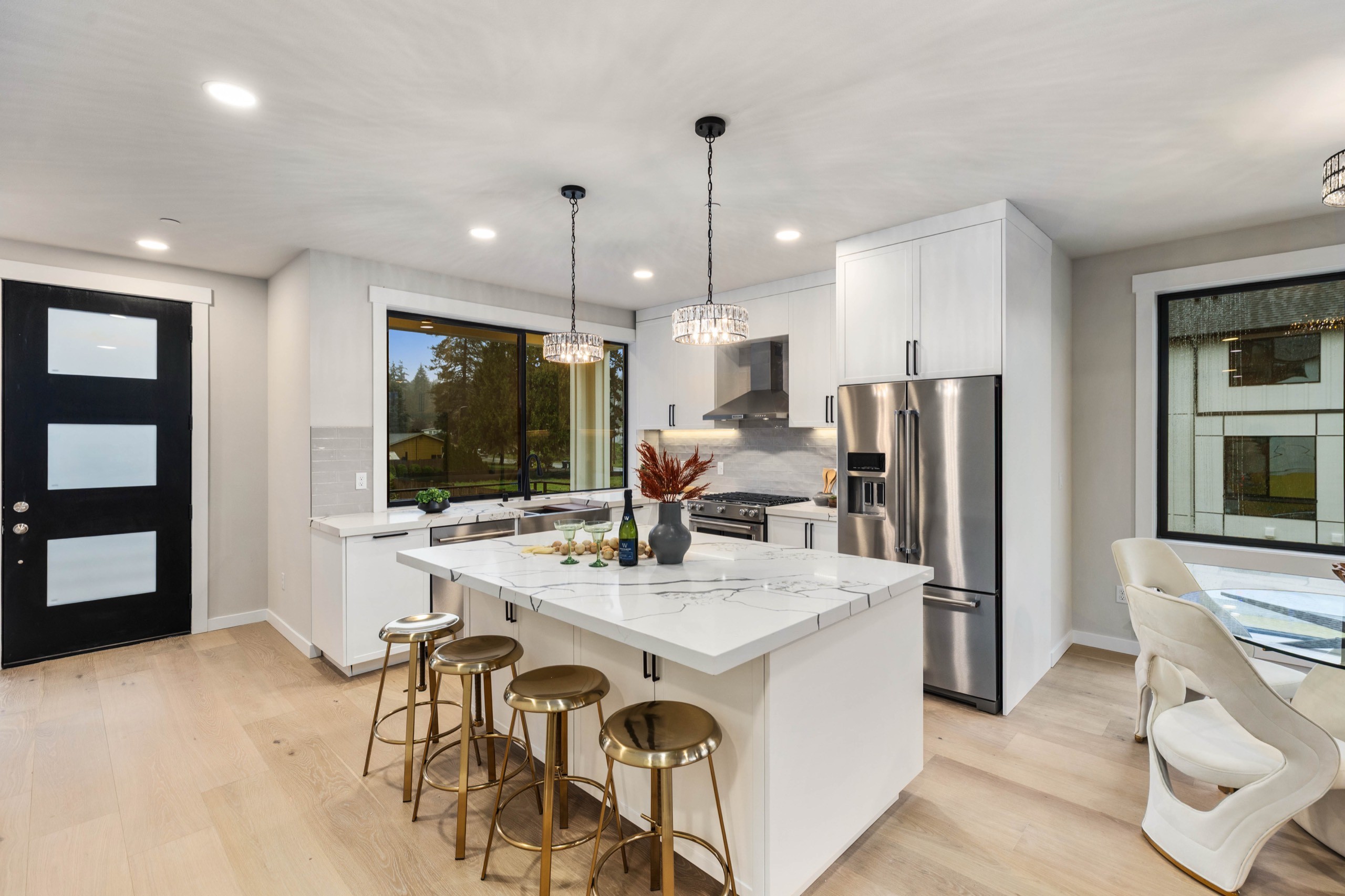 Bright luxury kitchen featuring white cabinetry, oversized island with gold pendant lights, stainless steel French door refrigerator, and black-framed glass entry door