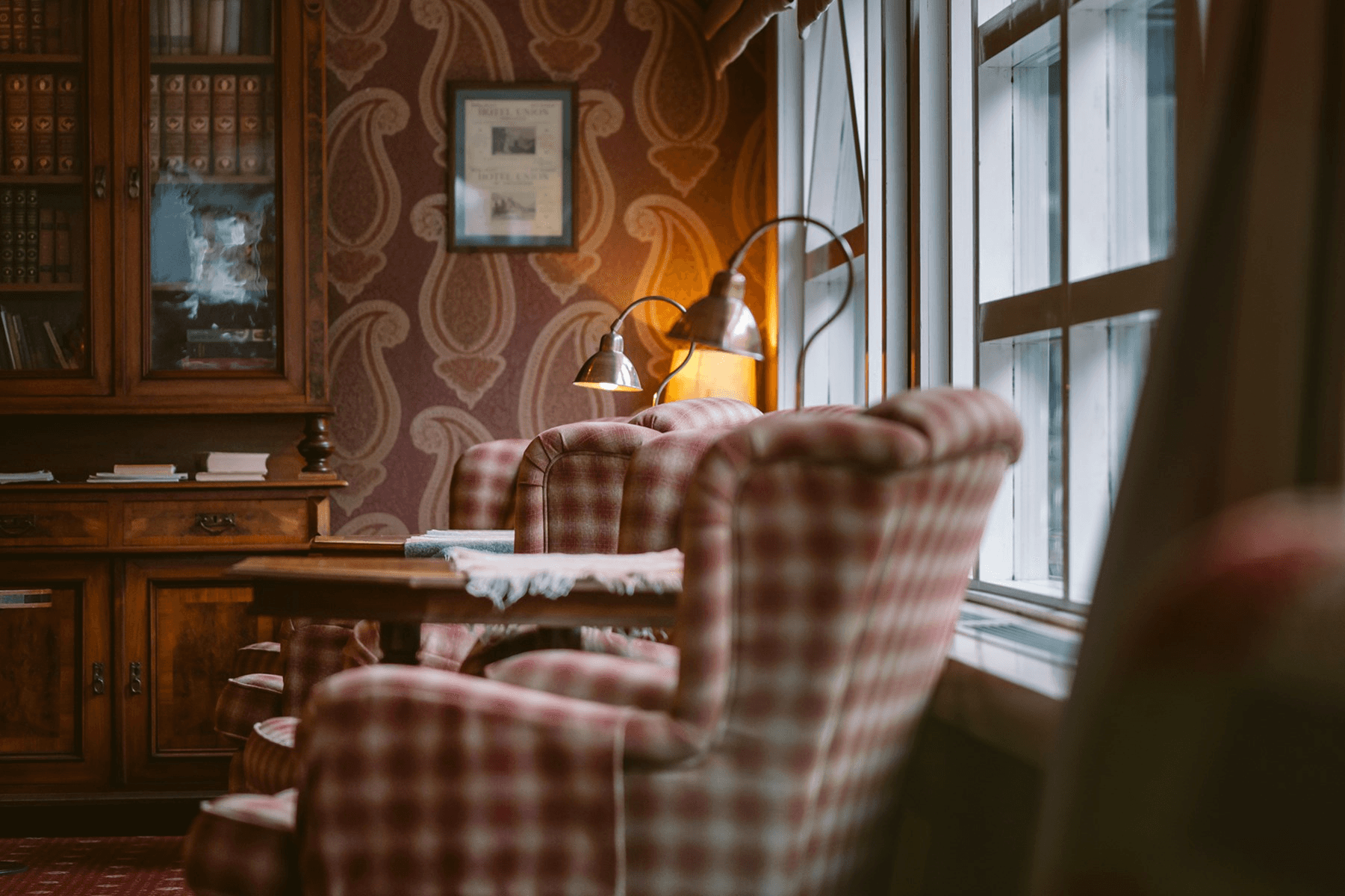 A cozy vintage library featuring plaid armchairs, a wooden desk, and a softly glowing lamp, beside a large window that invites natural light.