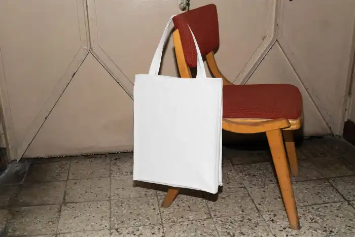 Canvas tote bag mockup in a muted pink shade, hanging from a vintage wooden chair with a red cushioned seat. The setting features a retro tiled floor and beige door, creating a nostalgic atmosphere.