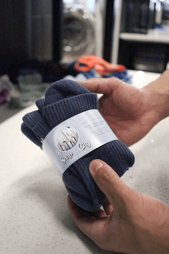 Hands holding neatly folded navy socks wrapped in a Soap and the City branded label, highlighting professional wash-and-fold laundry packaging and attention to detail.