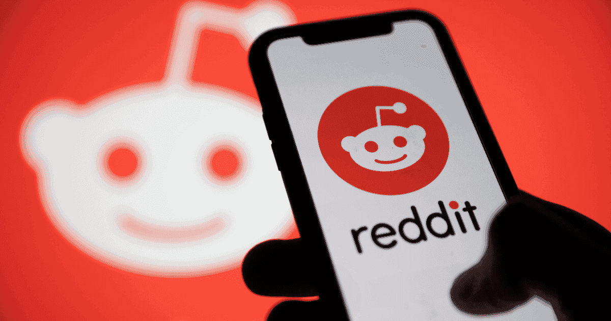 Reddit logo displayed on a phone screen held by a hand. The image is set against a red-blurry background carrying the head of a mascot in white, representing the Reddit logo. 