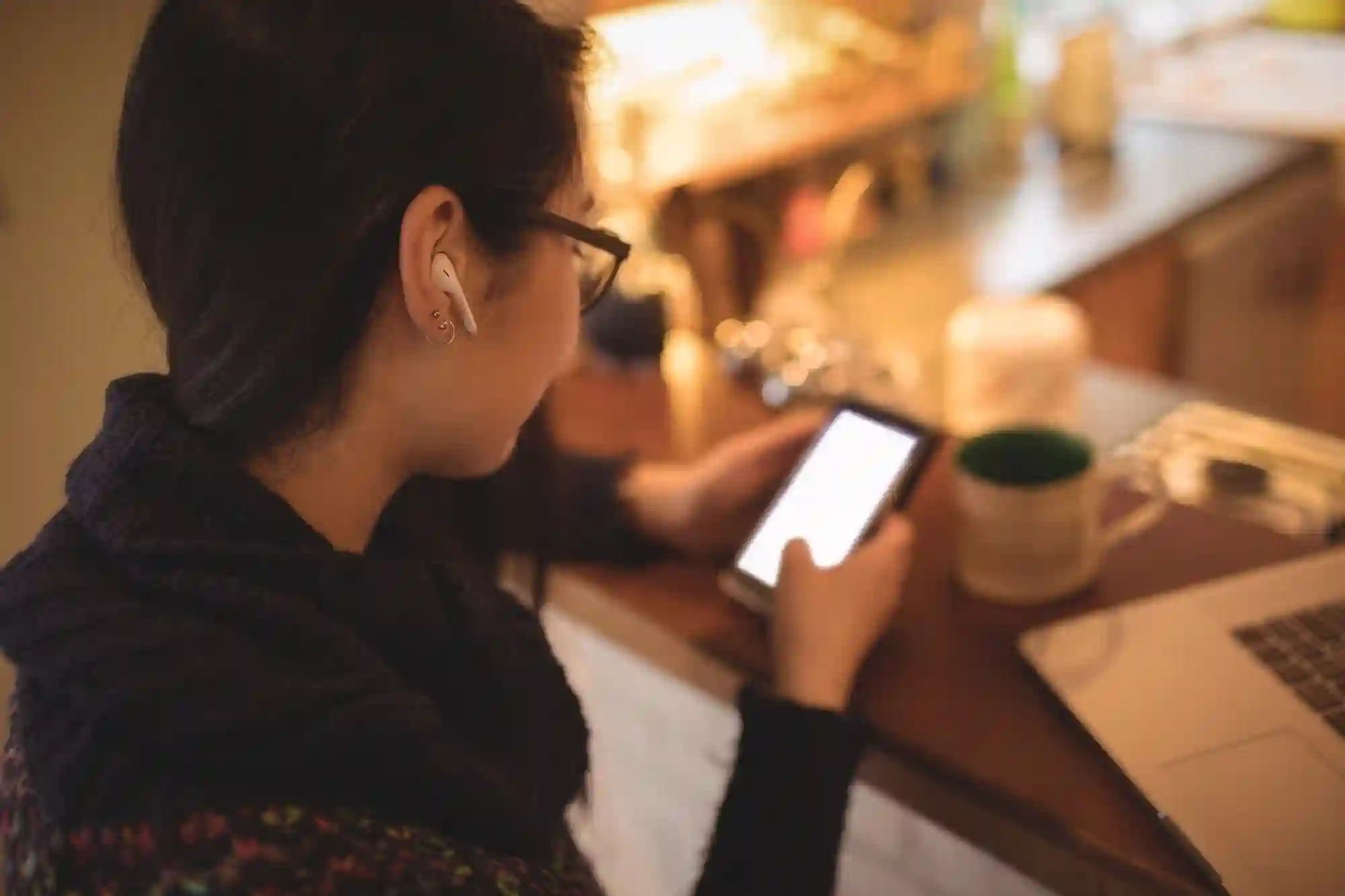 A woman using a smartphone and wireless earbuds while working at a desk with a laptop and coffee.
