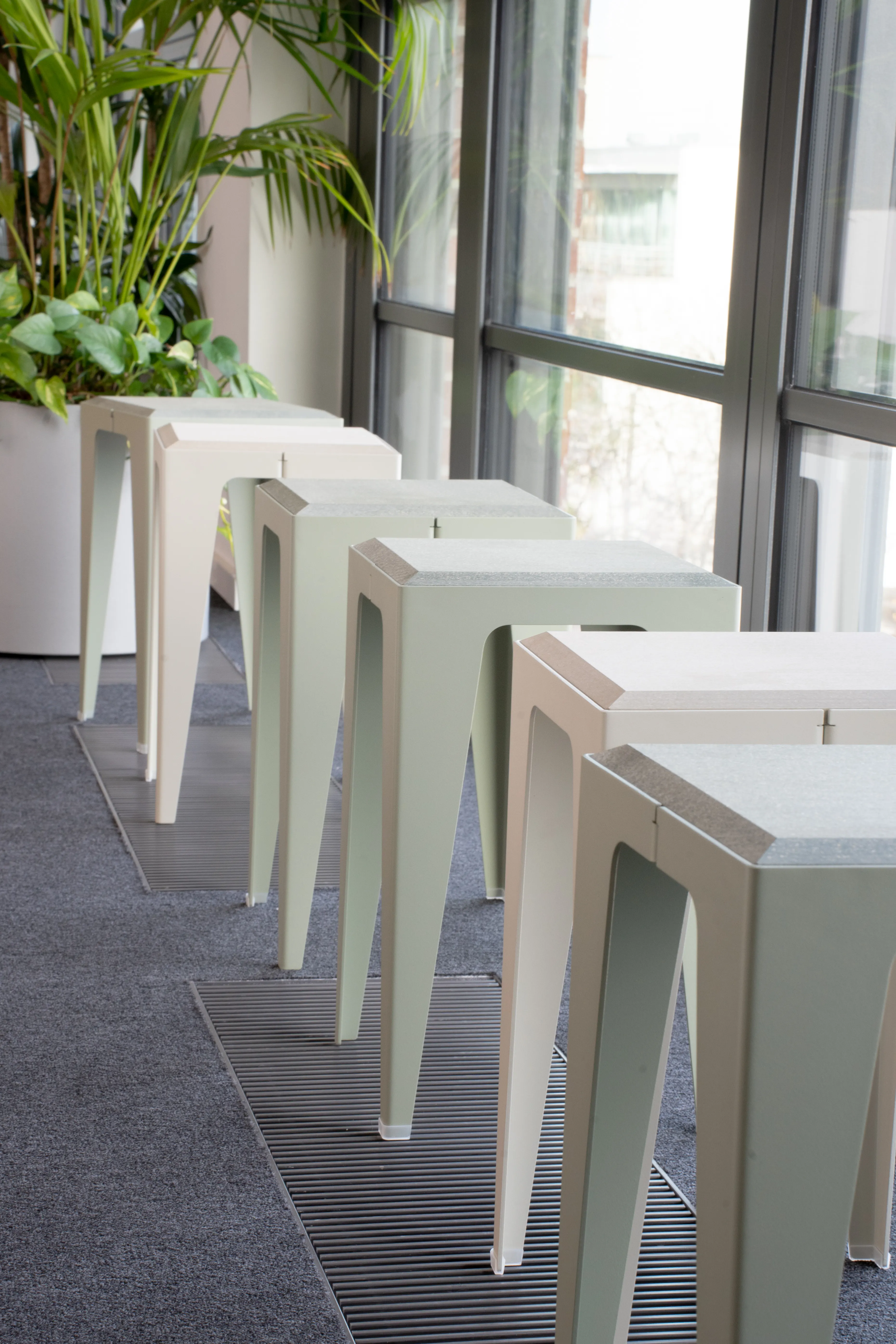 Row of angular stools in alternating cream and sage-green at AI START-UP MUNICH along a window front, with a large planter of palms and greenery behind.