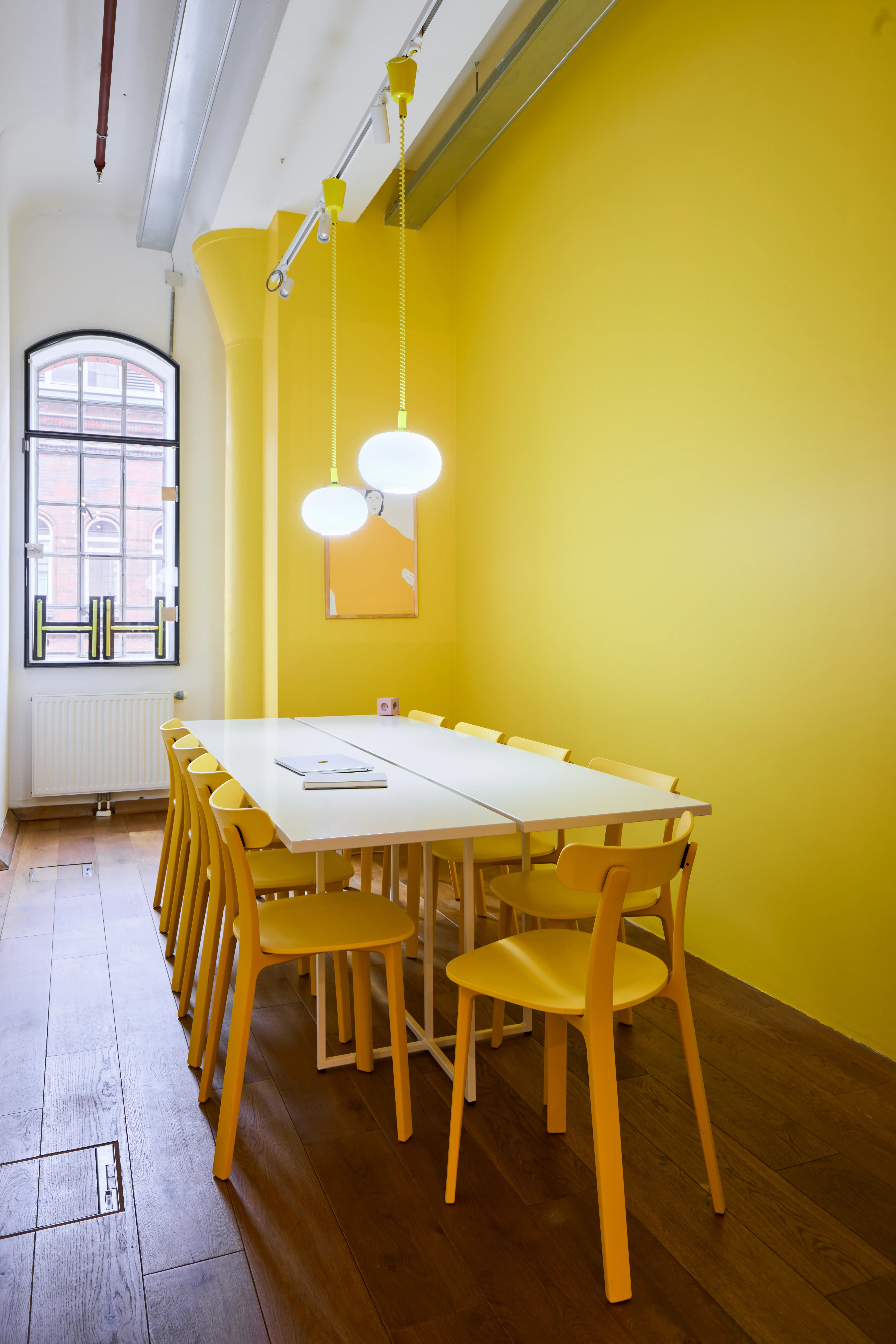 Yellow meeting room at INTERMATE OFFICE HAMBURG with a white table, yellow wooden chairs, two oval glass pendant lights, a yellow-painted column and wall, and an arched factory window.