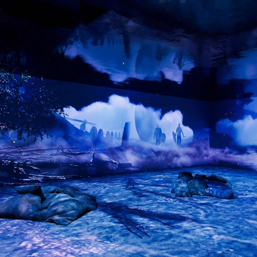 A room lit with blue, featuring a mural of a mystical landscape with silhouettes of people and giant moons. Pillows are scattered on the floor.
