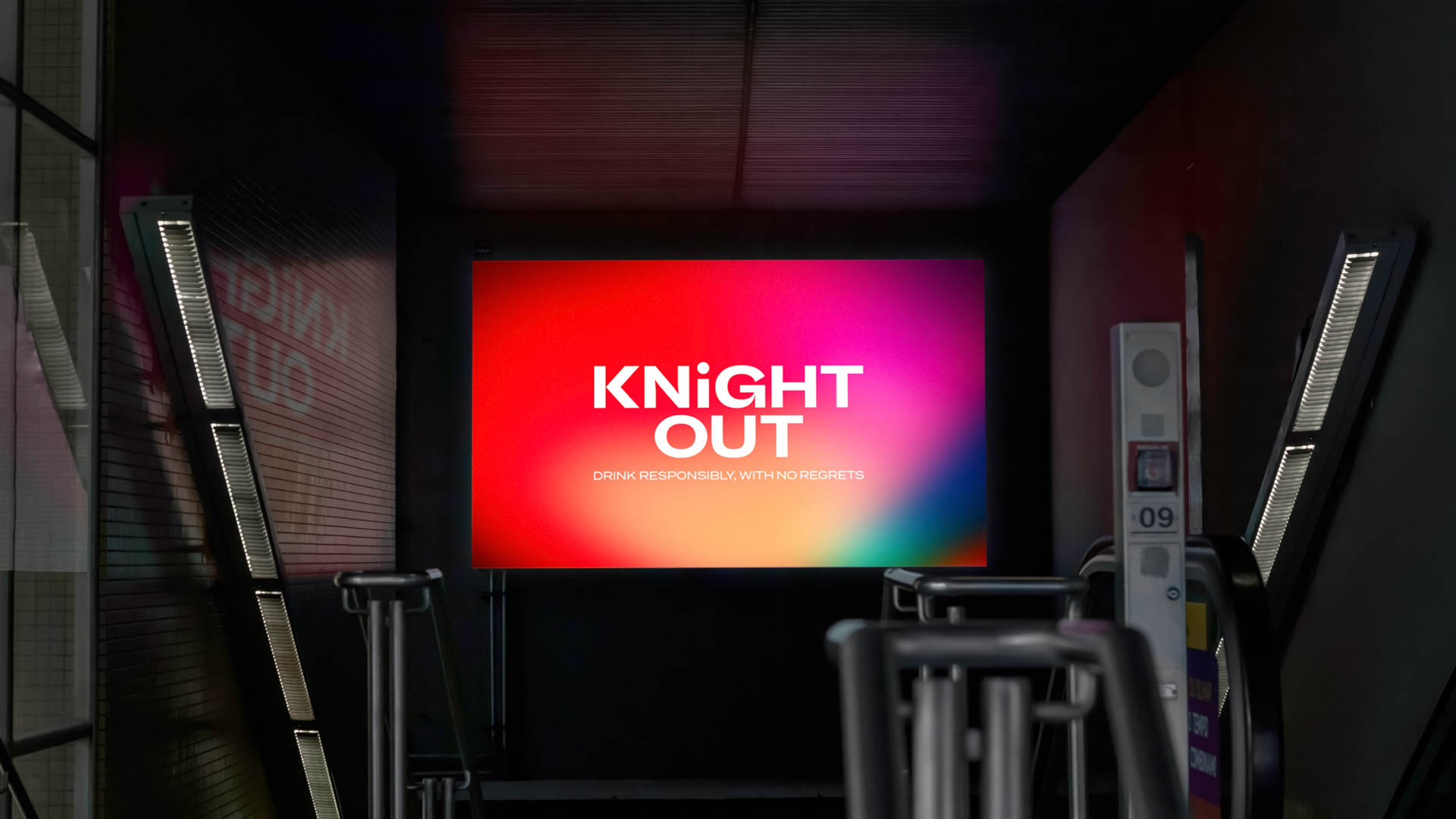 A large screen displays a red gradient background with the words "KNIGHT OUT" in white, centered. Below, smaller white text reads "DRINK RESPONSIBLY, WITH NO REGRETS." The screen is mounted on a dark wall with vertical metal panels on either side. Below the screen, a white device with the number "09" is visible.
