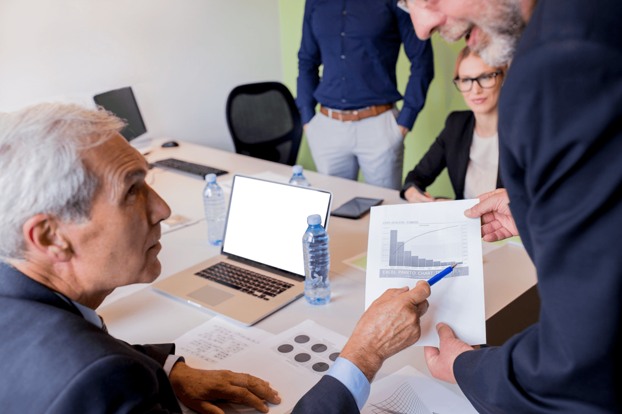 People in a meeting reviewing printed charts and data, with laptops and documents on the table.