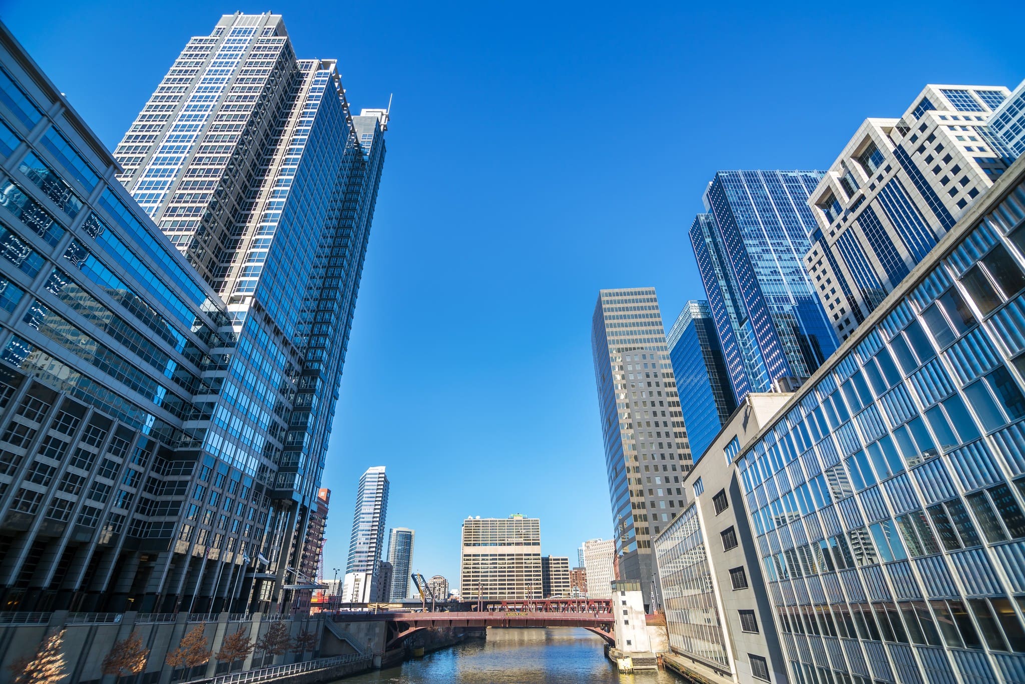 A low-angle view of an urban canal flanked by large commercial skyscrapers with bluish-gray glass facades, set against a clear sky. In the background, a metal bridge spans the water.