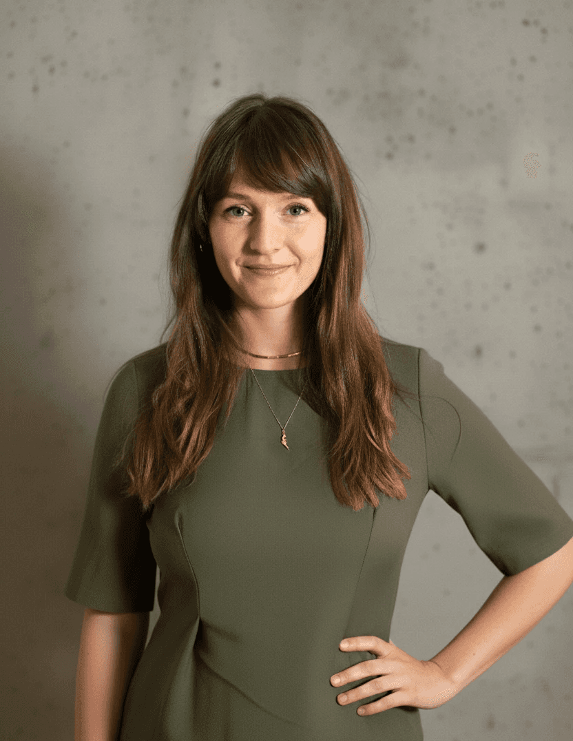 Photo of Nicole Zimmermann leaning agains a white wall, wearing a green blouse and slightly smiling in the camera