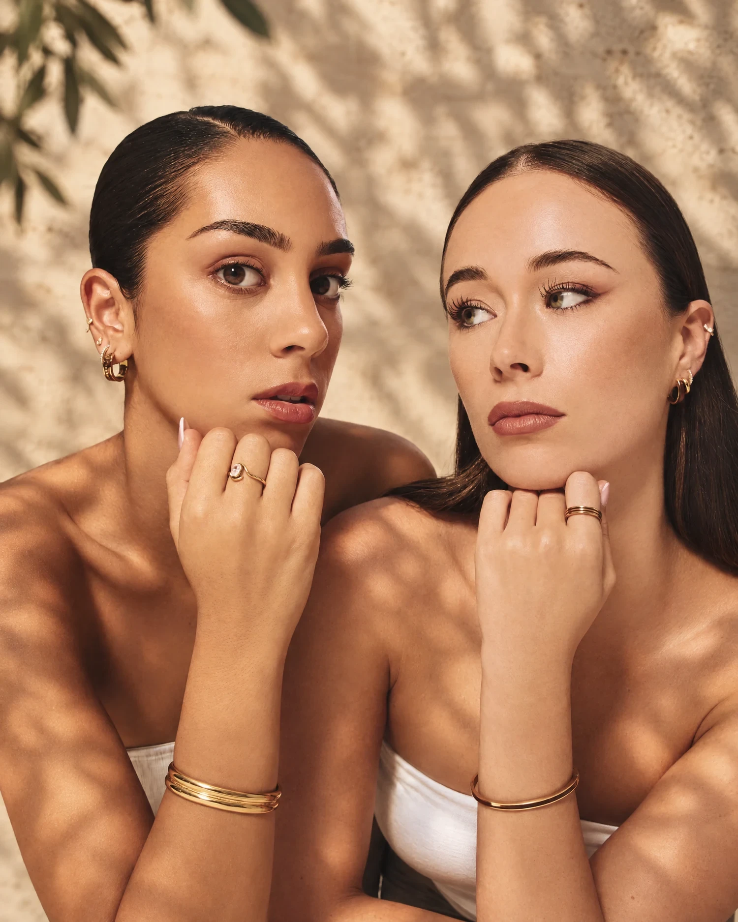 Two models from Kosmi's campaign in dappled afternoon sun against a cream travertine wall, chins on their hands, wearing gold cuffs, rings and earrings