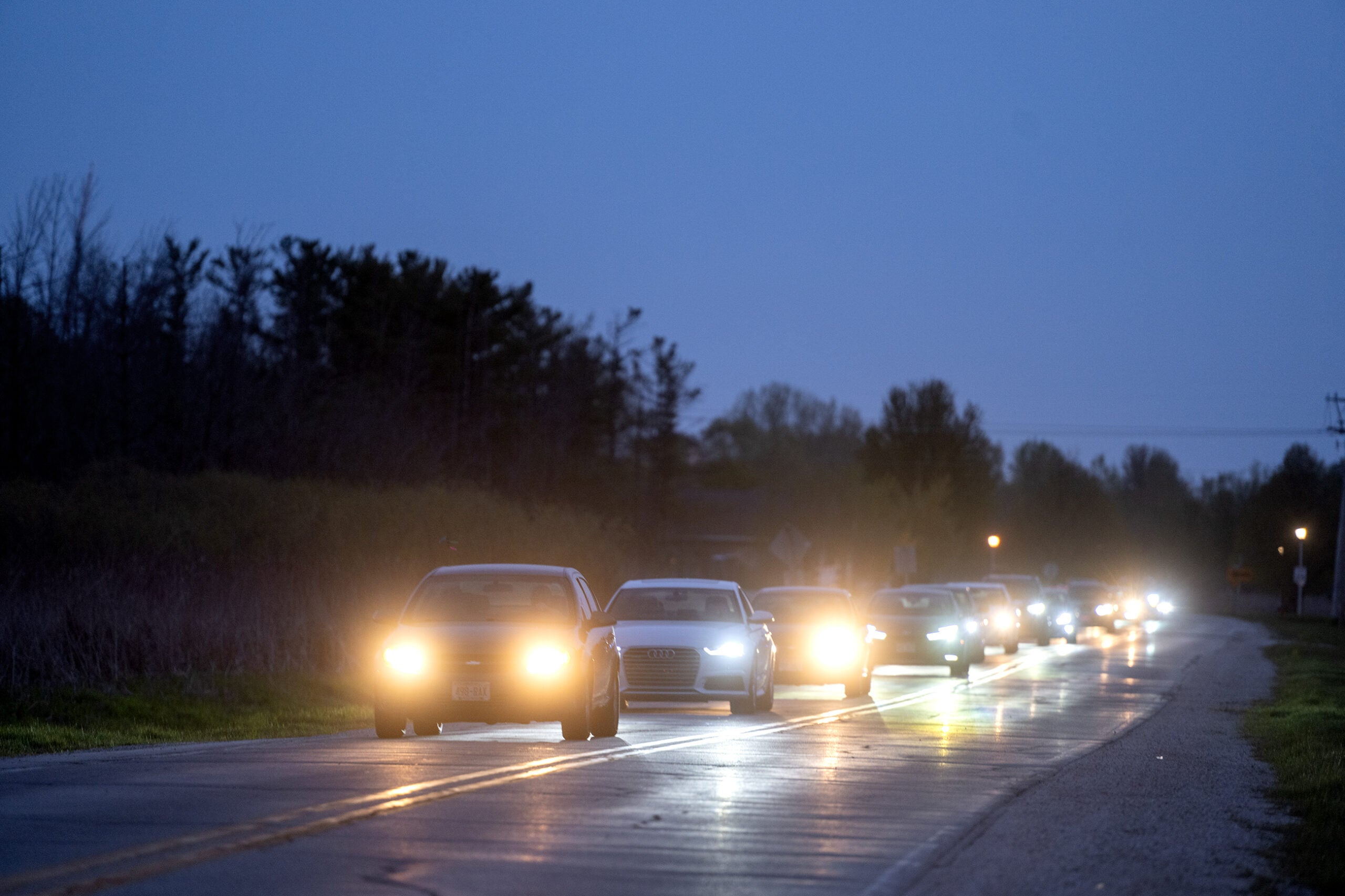 A line of cars with headlights on drives down a two-lane road at dusk, with trees and shrubbery visible along the sides.