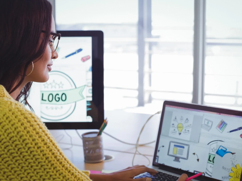 Graphic designer in yellow sweater working on a logo design at a desktop computer with dual screens