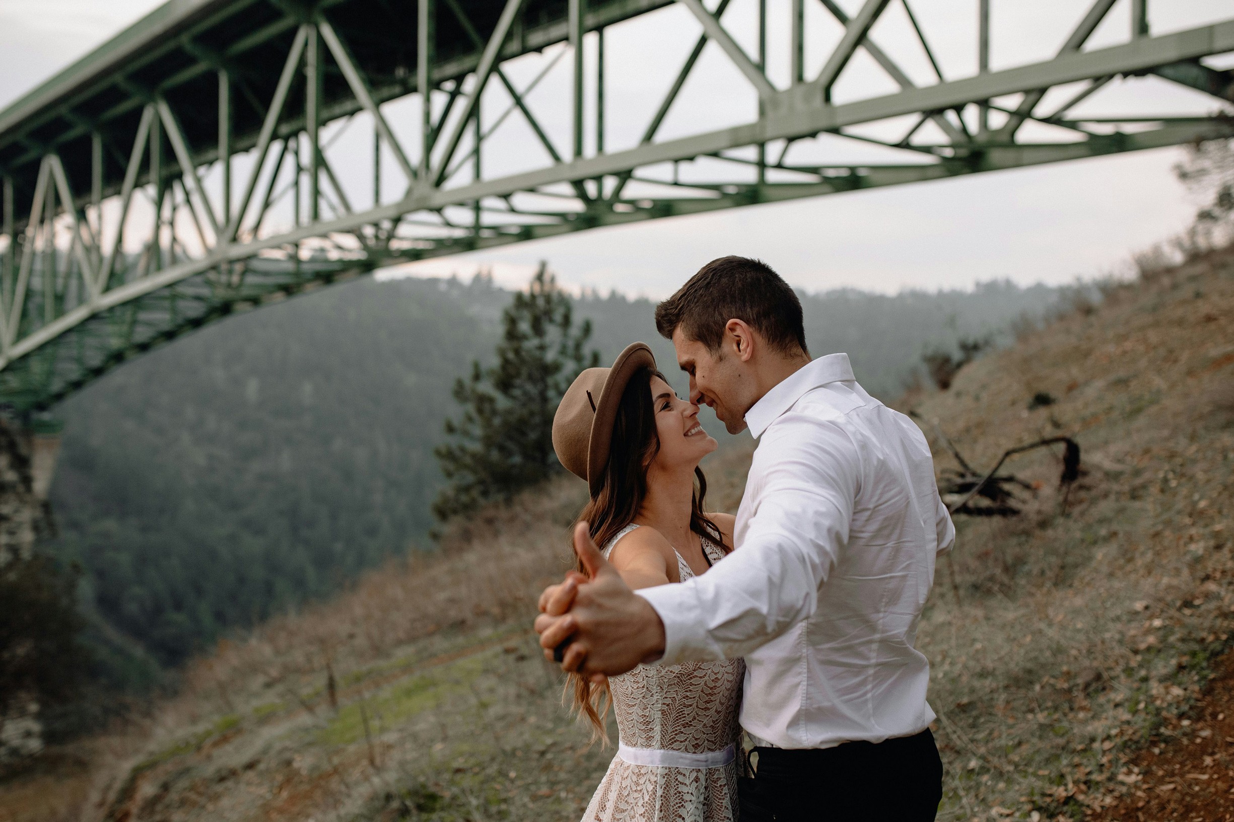 A couple embraces and smiles under a tall, green, steel bridge.