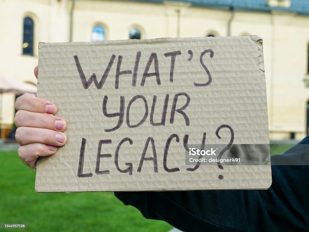 The image features a person holding a cardboard sign with the handwritten message, "WHAT'S YOUR LEGACY?" in bold letters. The background shows part of a building with arched windows, suggesting an outdoor setting, possibly a campus or public space. The sign's question prompts viewers to reflect on their impact and contributions, emphasizing themes of purpose and legacy.