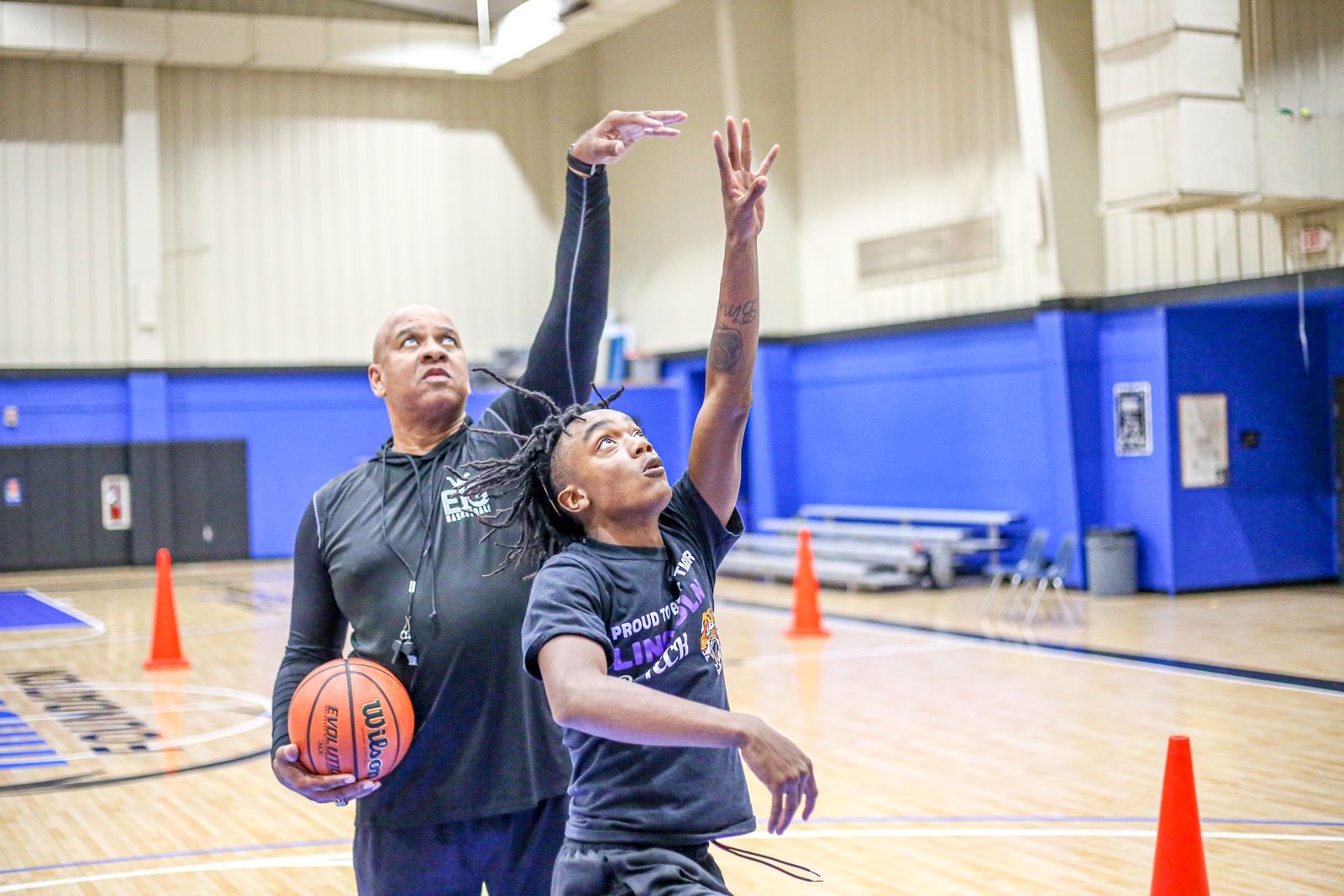 A coach guides a young player through a basketball shooting drill in a gym, focusing on technique.