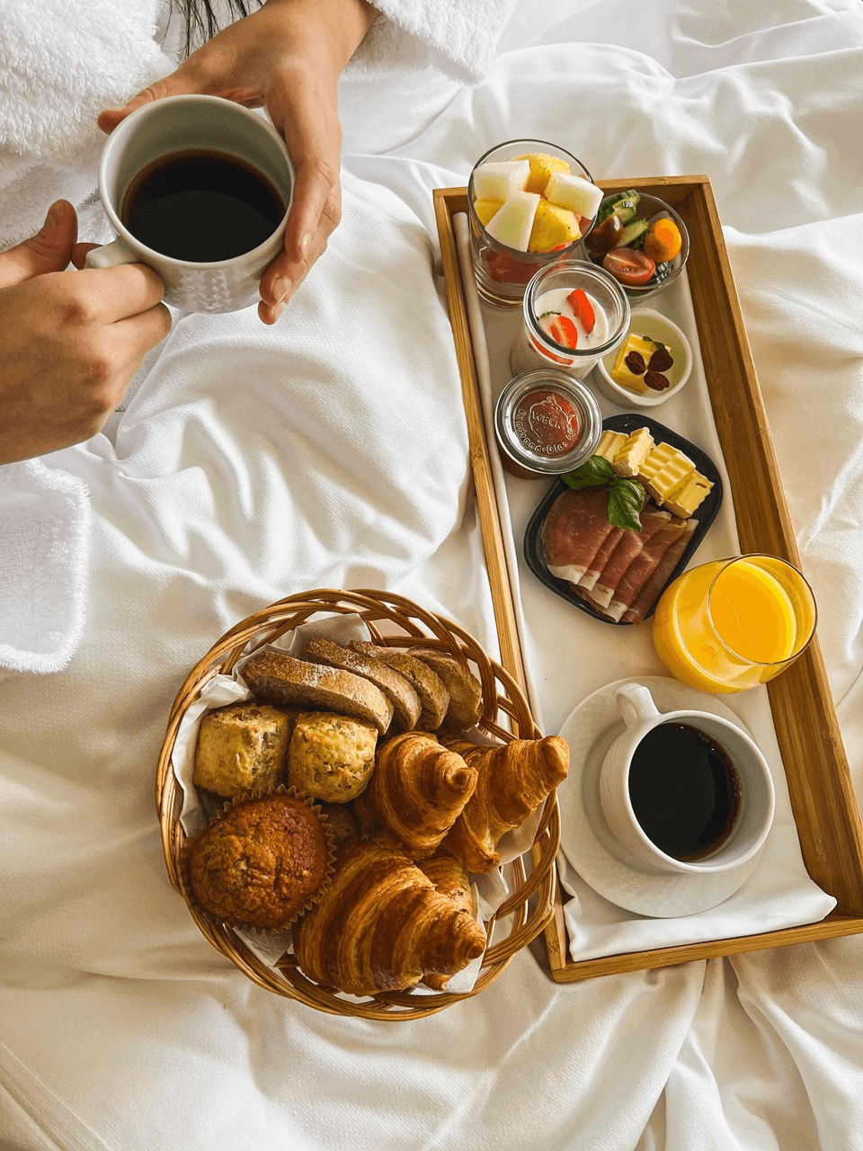 A breakfast in bed setup featuring a tray with sliced fruits, assorted cheeses, cured meats, a glass of orange juice, and a cup of coffee, alongside a basket of fresh pastries, including croissants and muffins.