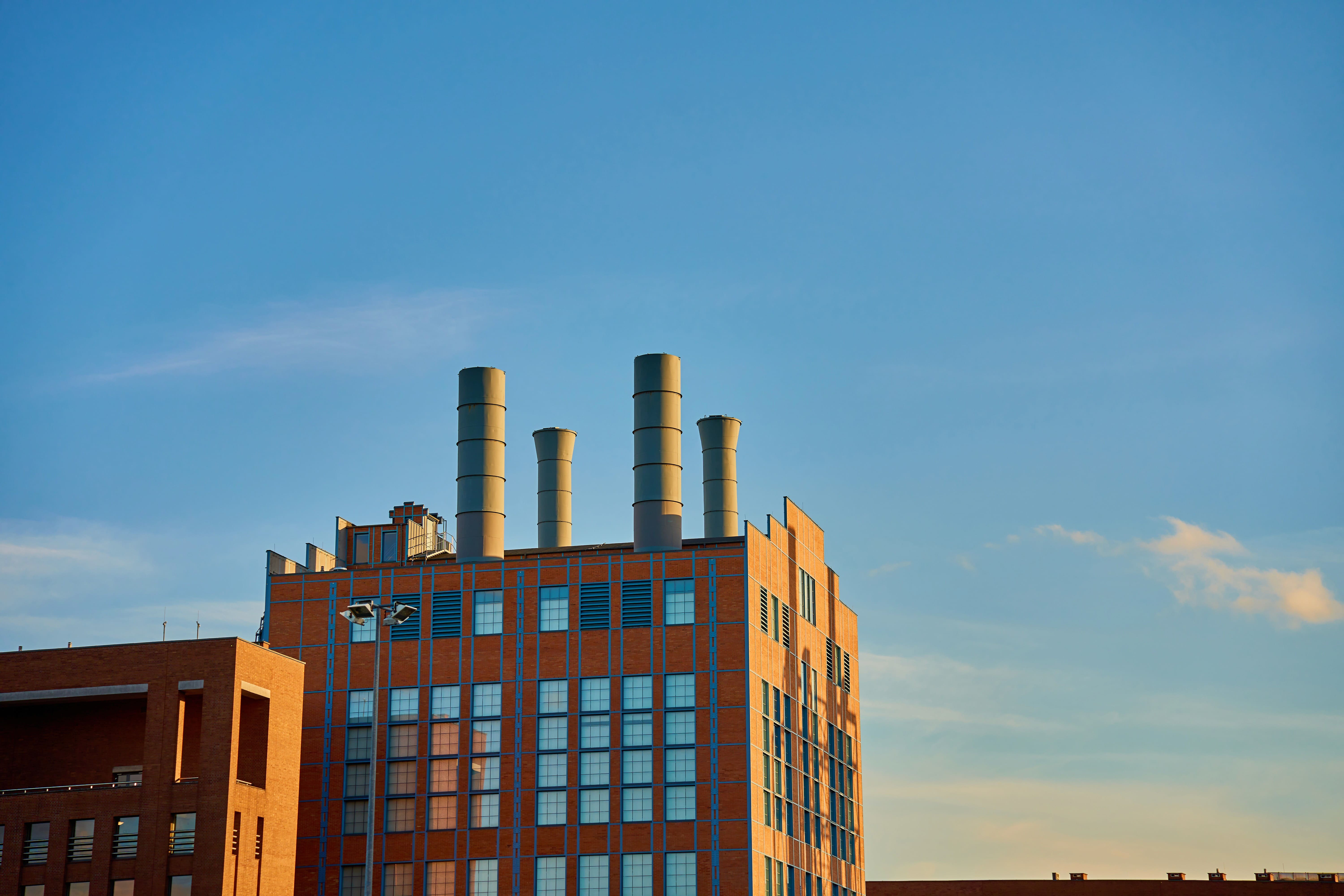 A brick industrial-style building featuring four tall grey exhaust chimneys on its roof, illuminated by the warm light of late afternoon against a clear blue sky.