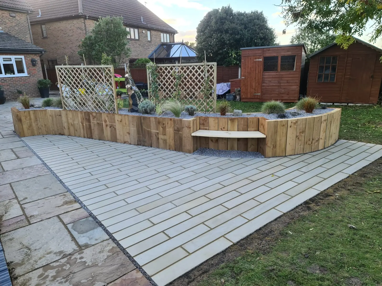 A landscaped patio area featuring wooden planters, a wooden bench, and pavers, surrounded by green grass.