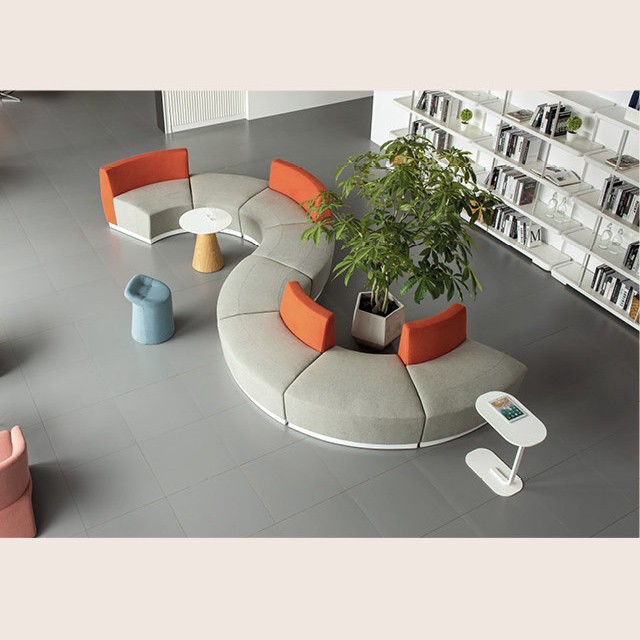 Modern S-shaped modular sectional sofa in gray and orange fabric, arranged in a contemporary office lobby with a large potted plant and minimalist white side tables.