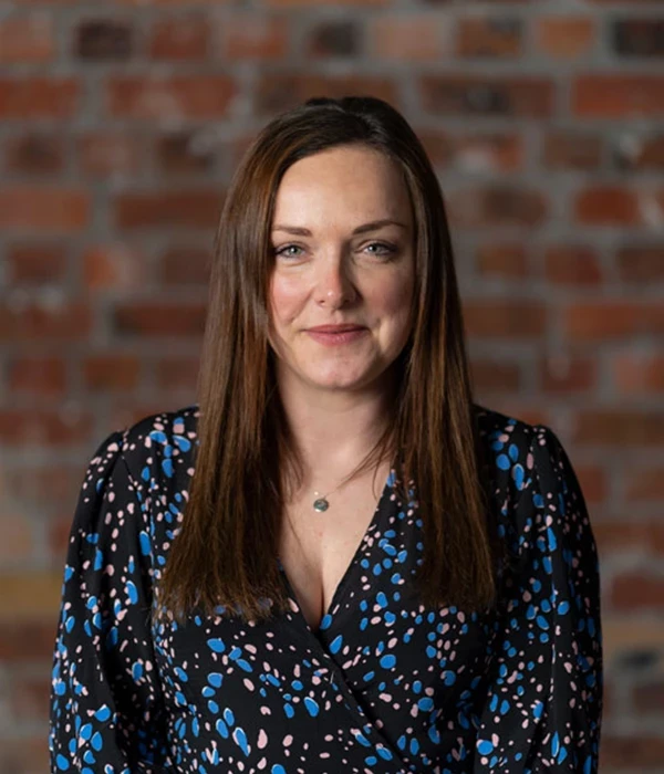 A portrait‑style image of a person shown from the chest up, standing against an exposed red‑brick wall. They are wearing a dark blouse patterned with small blue, white, and pink abstract shapes and a delicate necklace with a small pendant. Their straight, medium‑length brown hair falls neatly over their shoulders. The lighting is soft and even, giving the image a clean, professional feel suitable for a profile or team photograph.