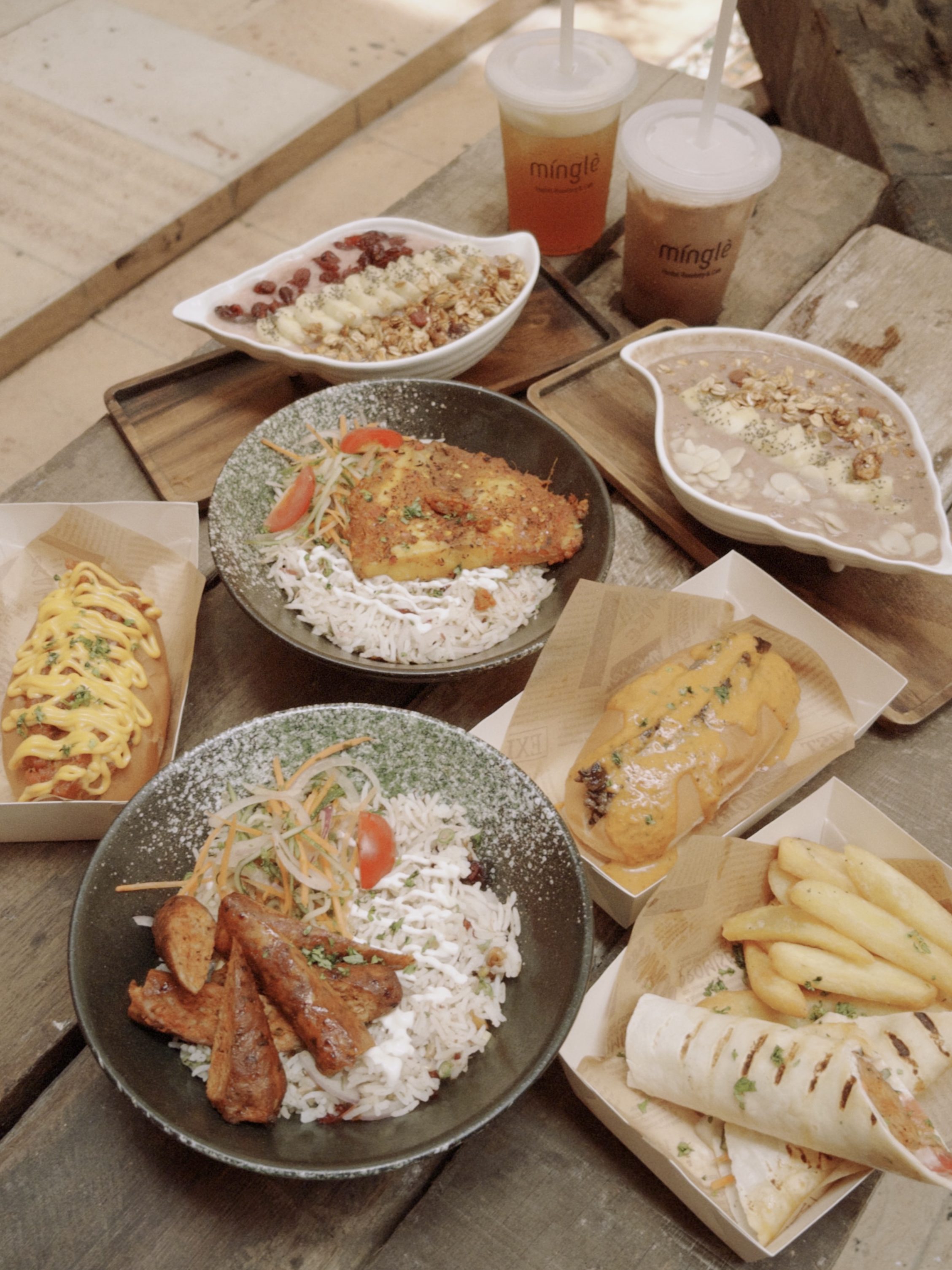 A spread of Malaysian and Western dishes served on a rustic wooden table, featuring rice, fried chicken, hot dogs, and beverages from Liku Likoo Lekiu café.