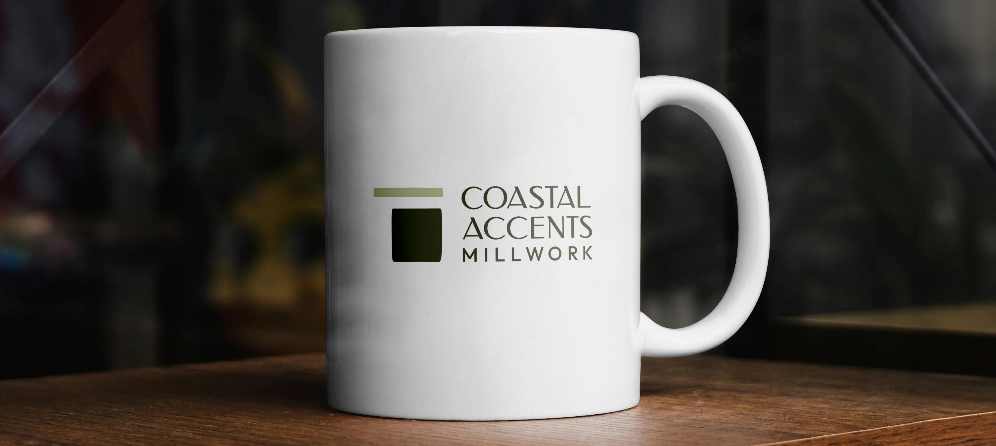 A white mug on a wooden table with the Coastal Accents logo on it.
