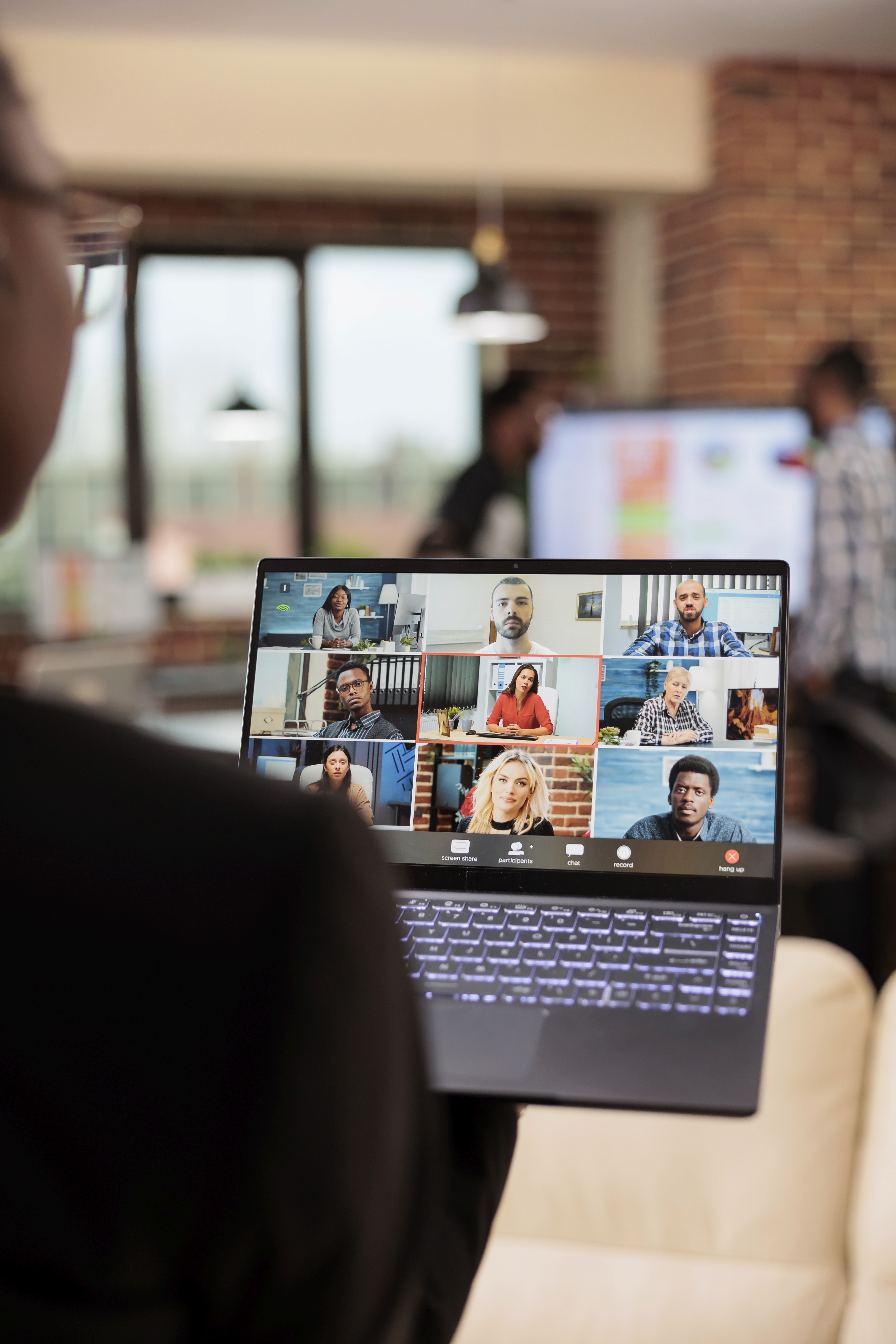 Eine Person steht in einem Büro. Sie hält einen Laptop in der Hand, der groß im Bild zu sehen ist. Die Person befindet sich in einem Video-Call mit anderen Teammitgliedern, die auf den Bildschirm zu sehen sind.