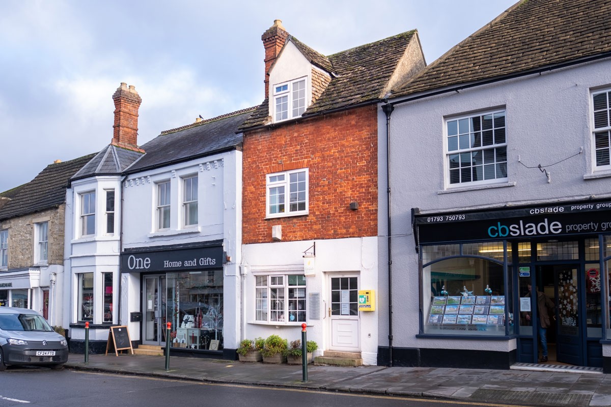 A picturesque streetscape featuring a row of charming, traditional British shops with brick and white facades, including Cricklade Dental Practice situated on a calm, overcast day.