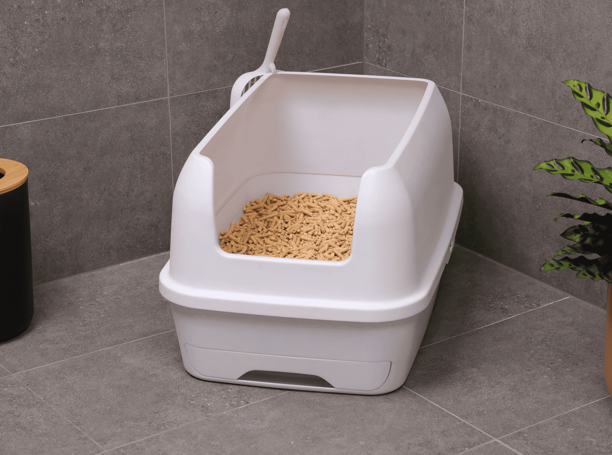 A container of sand-like cat litter is being poured into a lined plastic trash bin next to a modern, white litter box on a tiled bathroom floor with a leafy, potted plant nearby.