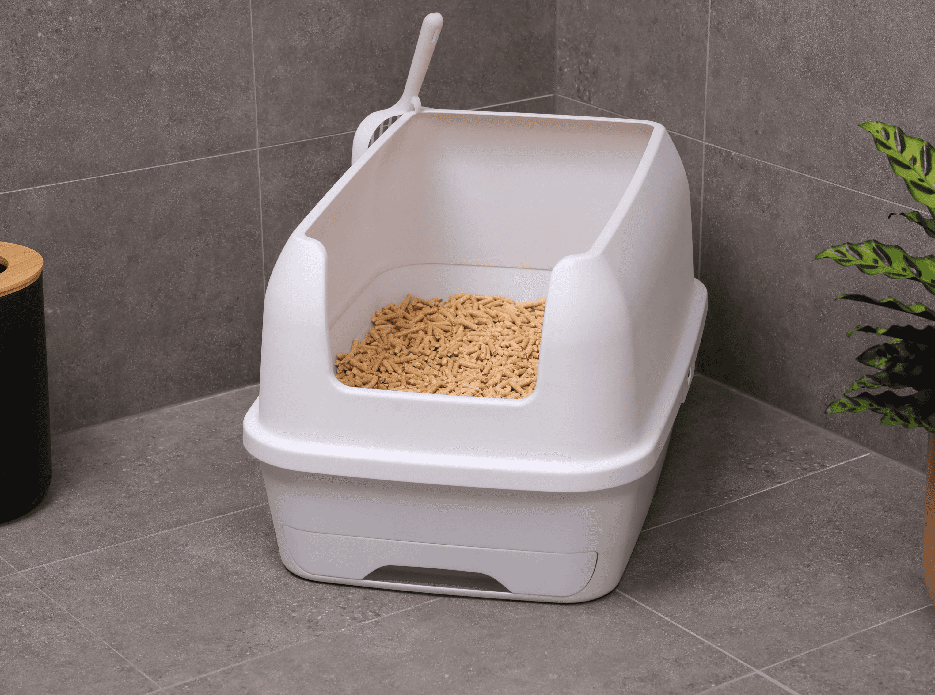 A container of sand-like cat litter is being poured into a lined plastic trash bin next to a modern, white litter box on a tiled bathroom floor with a leafy, potted plant nearby.