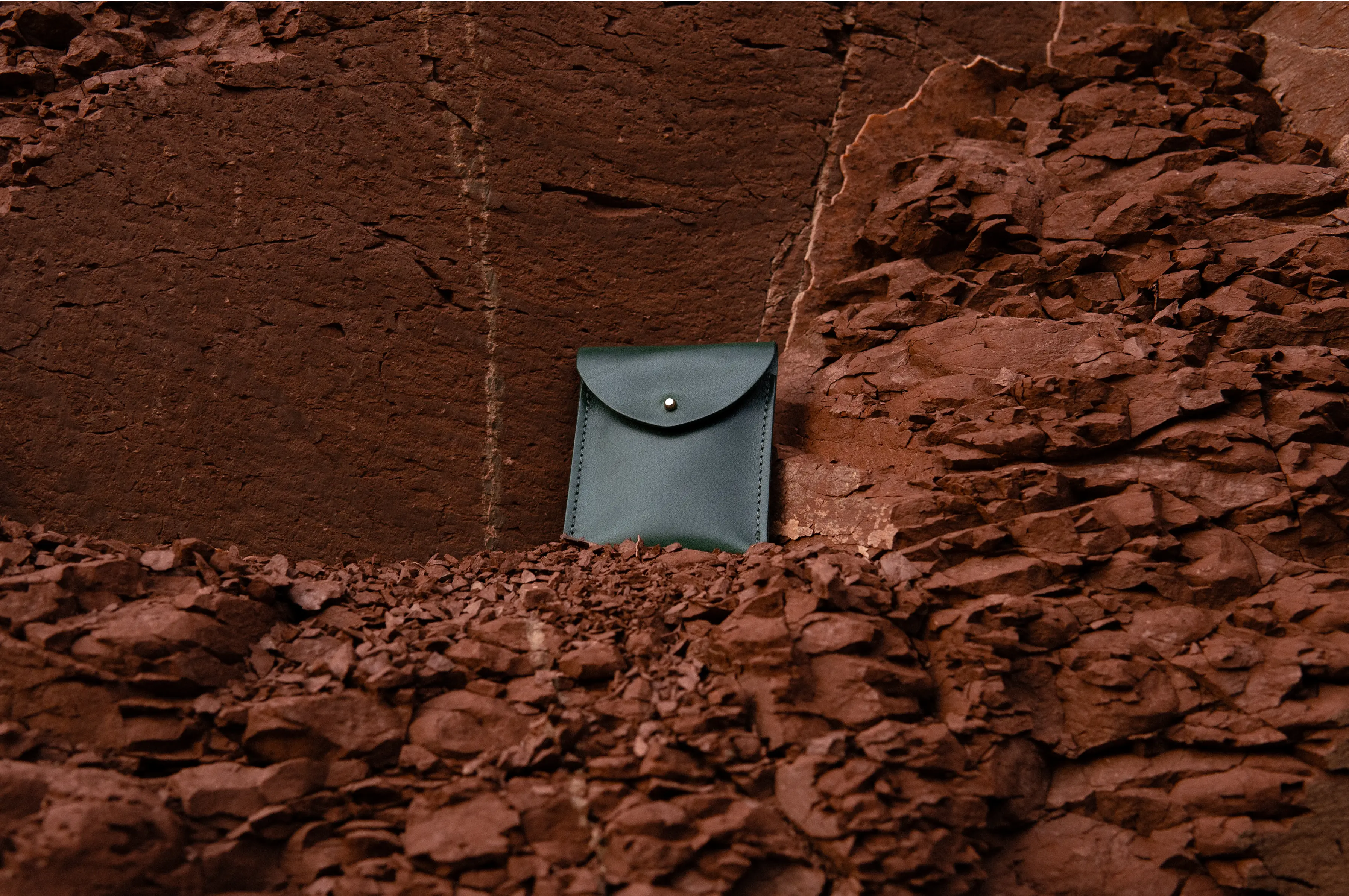 Small dark green leather pouch placed between red rocks, photographed as a fashion campaign object.