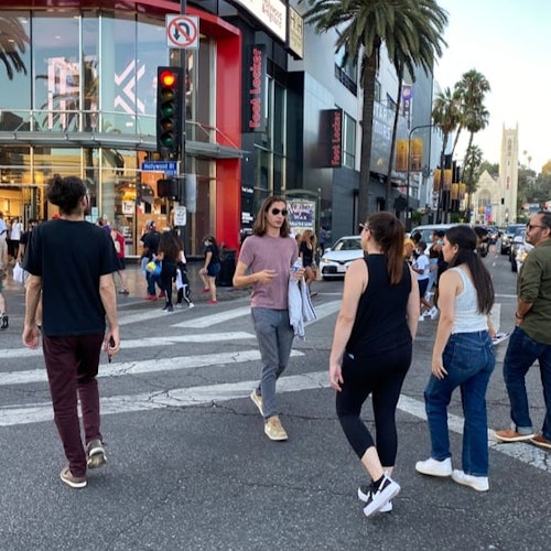 People crossing a busy city street with shops, palm trees, and a church in the background.