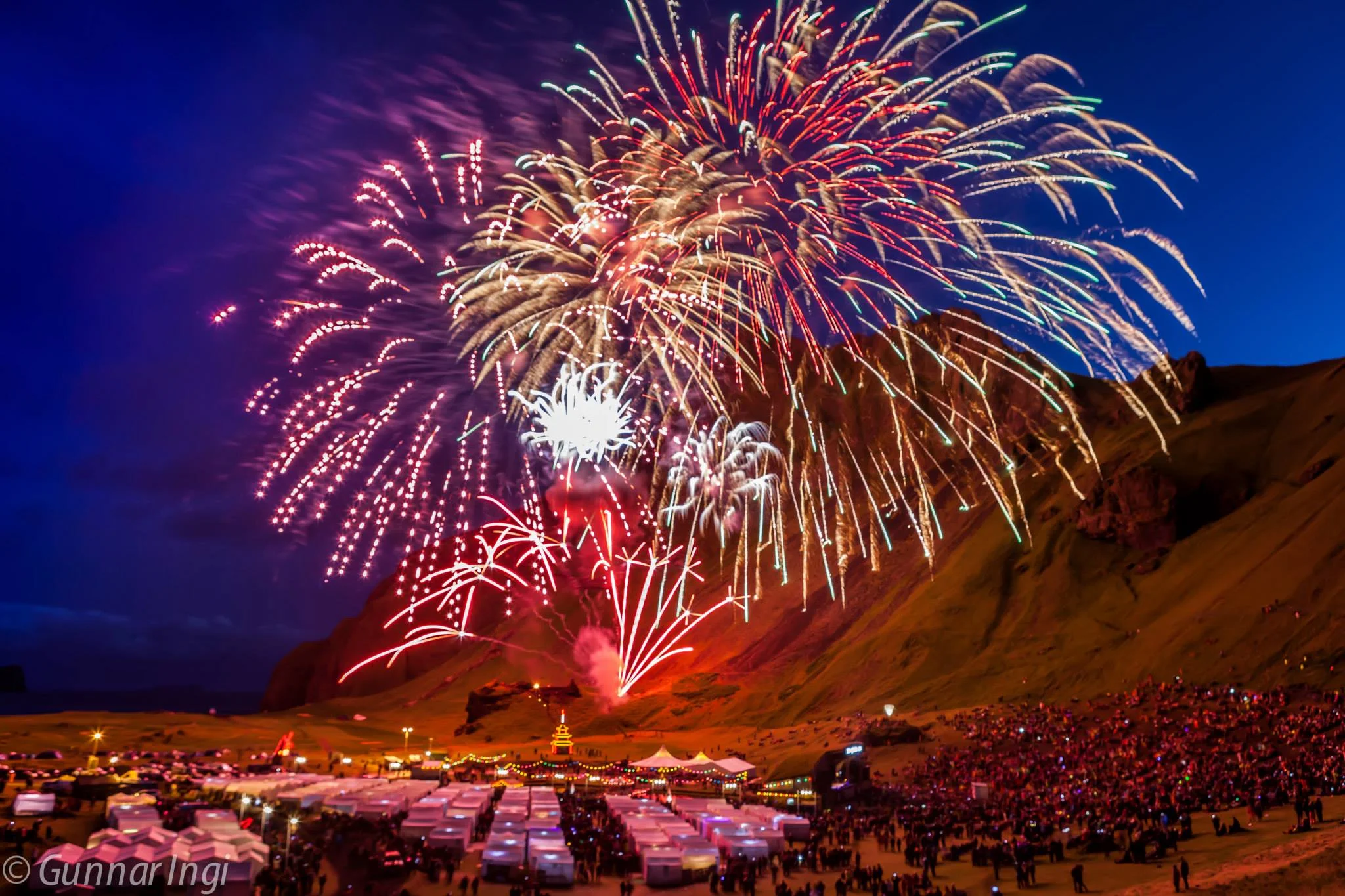 A massive fireworks display illuminates the night sky in vibrant colors over a crowded festival valley in the Westman Islands Herjólfsdalur Valley on Heimaey Island with rows of white tents beneath a large volcanic hill.
