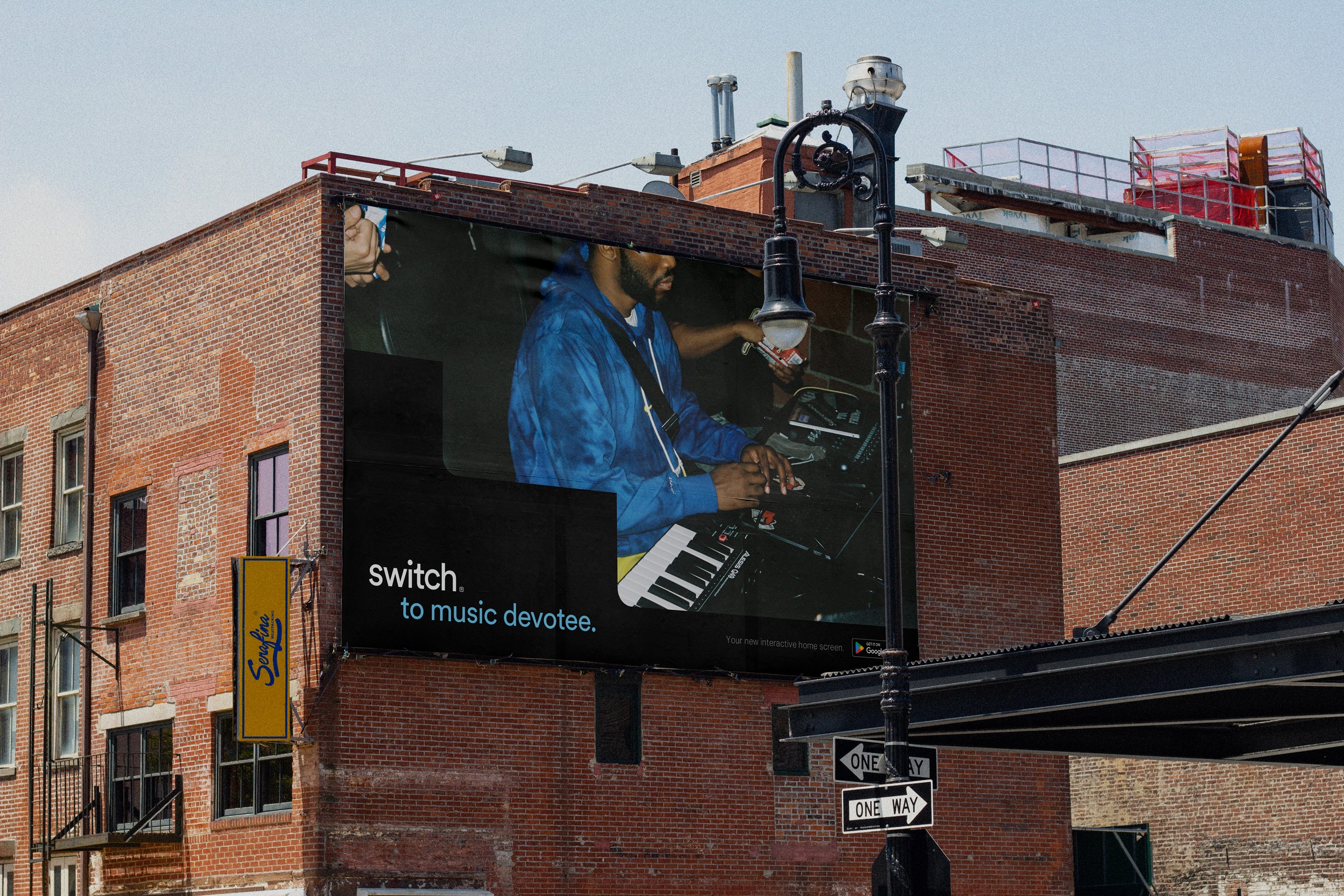 A large billboard on the side of a red brick building displays a musician playing a keyboard. The billboard features the text "switch. to music devotee." A street sign reading "ONE WAY" and a street lamp are visible below the billboard.