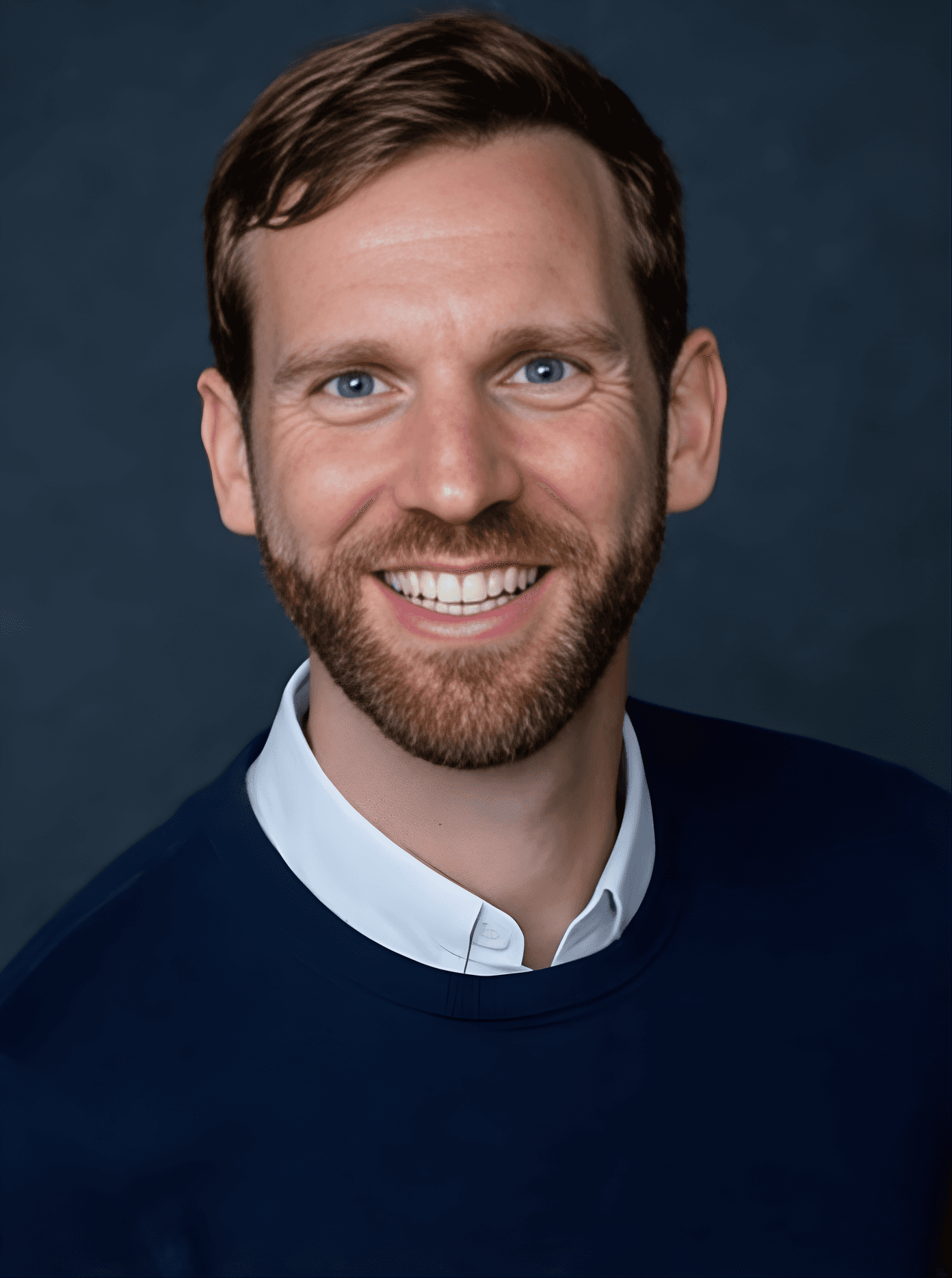 A smiling person with curly hair, wearing a leather jacket, stands in a cozy indoor setting.