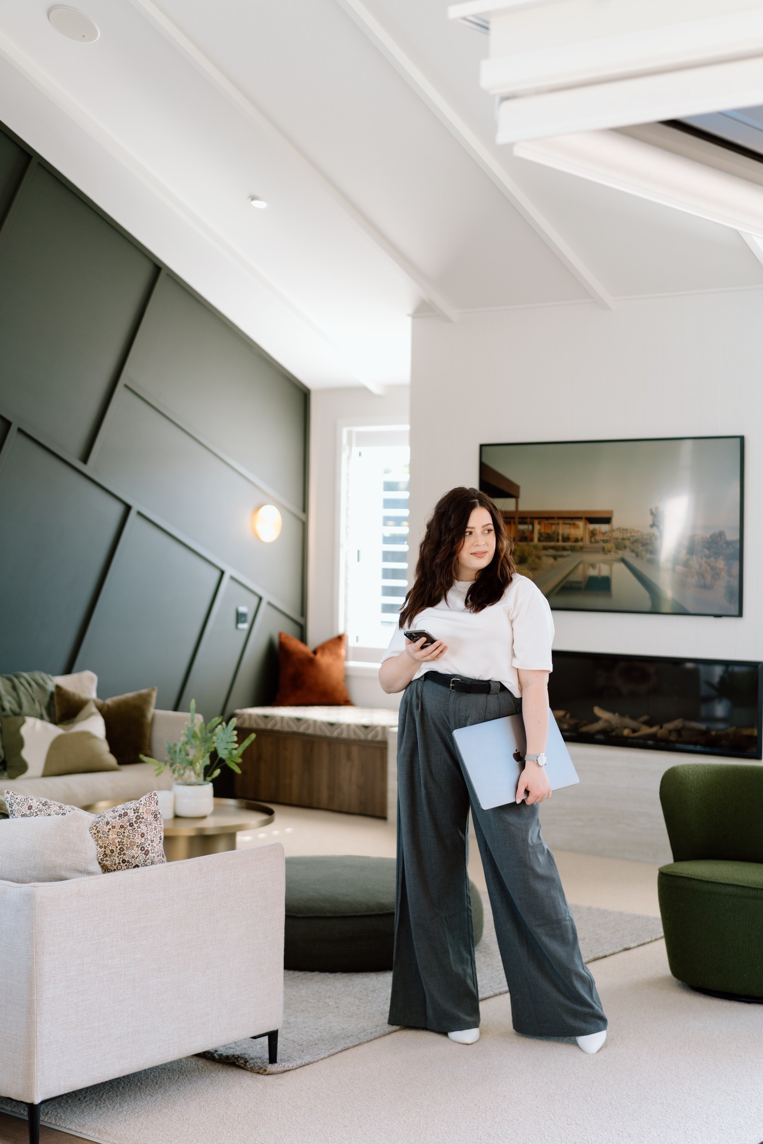 A woman in grey professional pants and white tshirt holding a computer, standing amongst couches in a lounge area