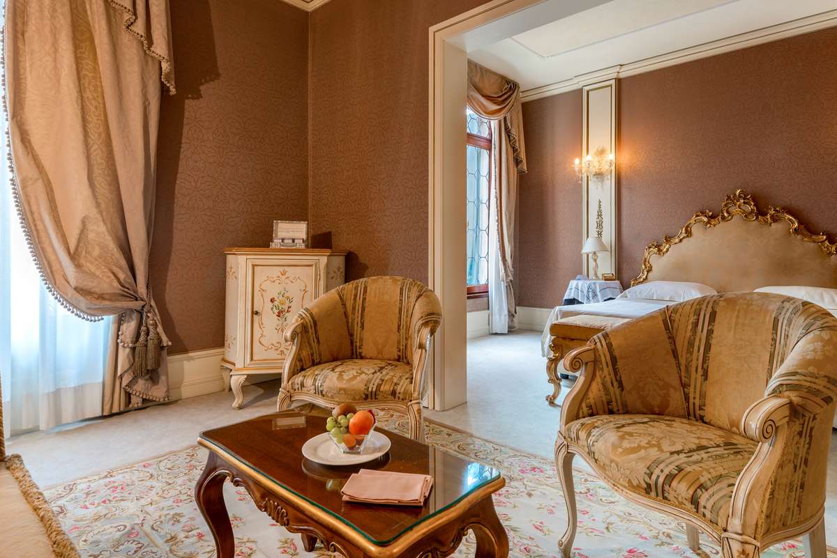 Two ornate gold and beige armchairs face a wooden table with fruit and books in a bedroom with brown walls and drapes.