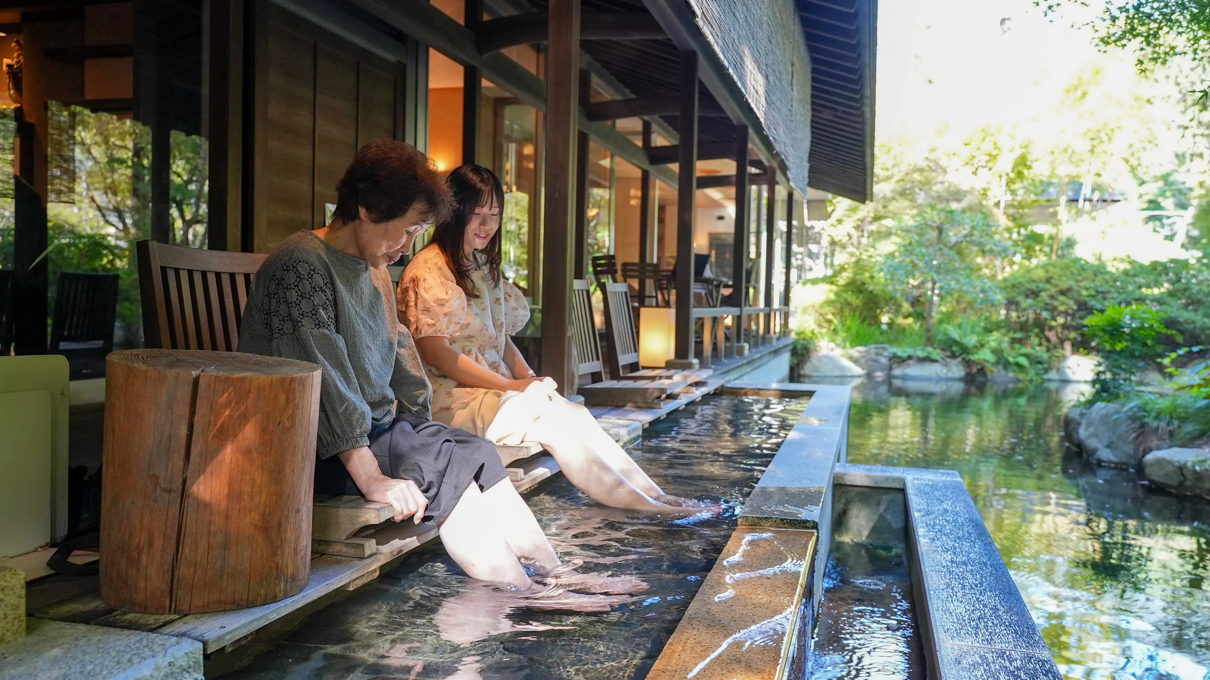Two families enjoy a relaxing foot bath (ashiyu) on a wooden deck that overlooks a large pond filled with colorful koi fish.