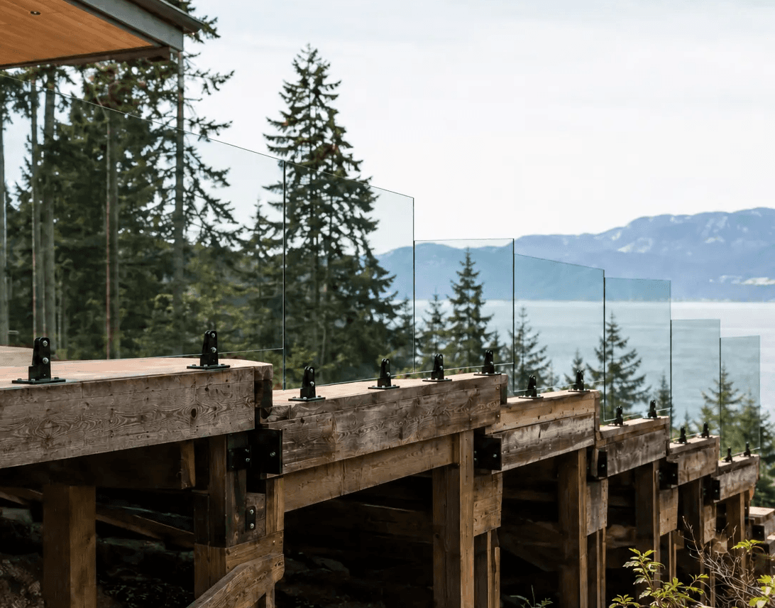 Modern cedar deck with frameless glass railings and black mounting hardware overlooking a forested lakeside mountain view. Exposed old-growth timber construction and warm cedar soffits create a clean west coast contemporary design.