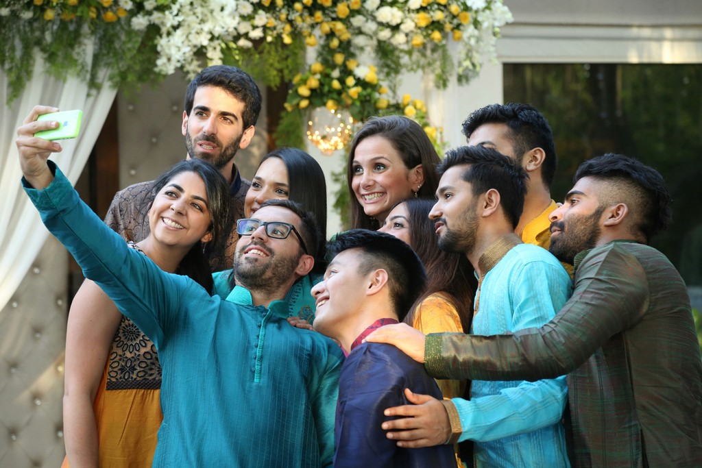 A diverse group of friends in colorful traditional attire gather closely to take a joyful selfie, celebrating together at a festive outdoor event adorned with flowers and decorations.