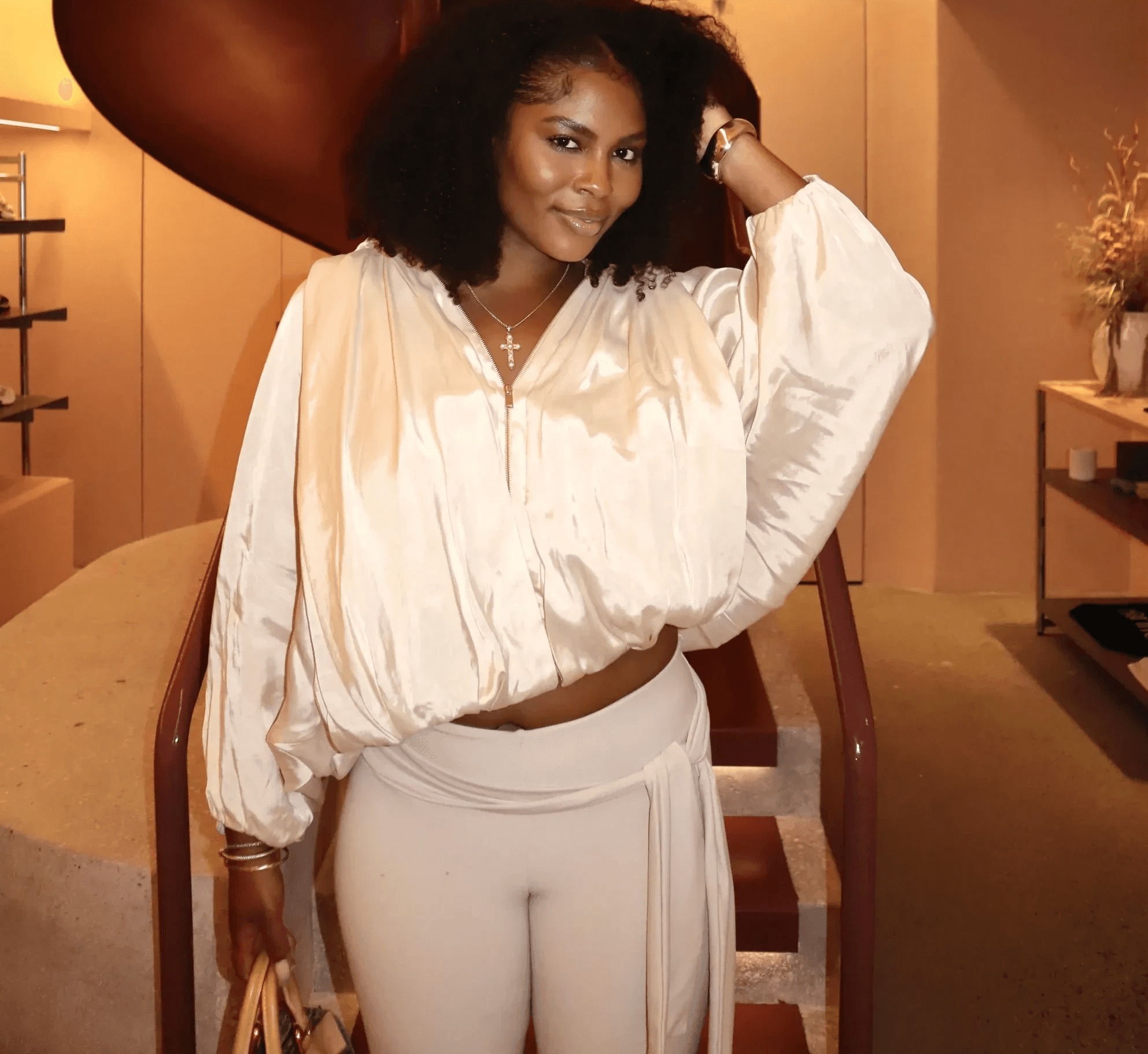 Dr. Micah Brown with curly hair, wearing a loose cream blouse and matching pants, stands confidently in front of a modern, dark red spiral staircase in a stylish, warmly lit interior.